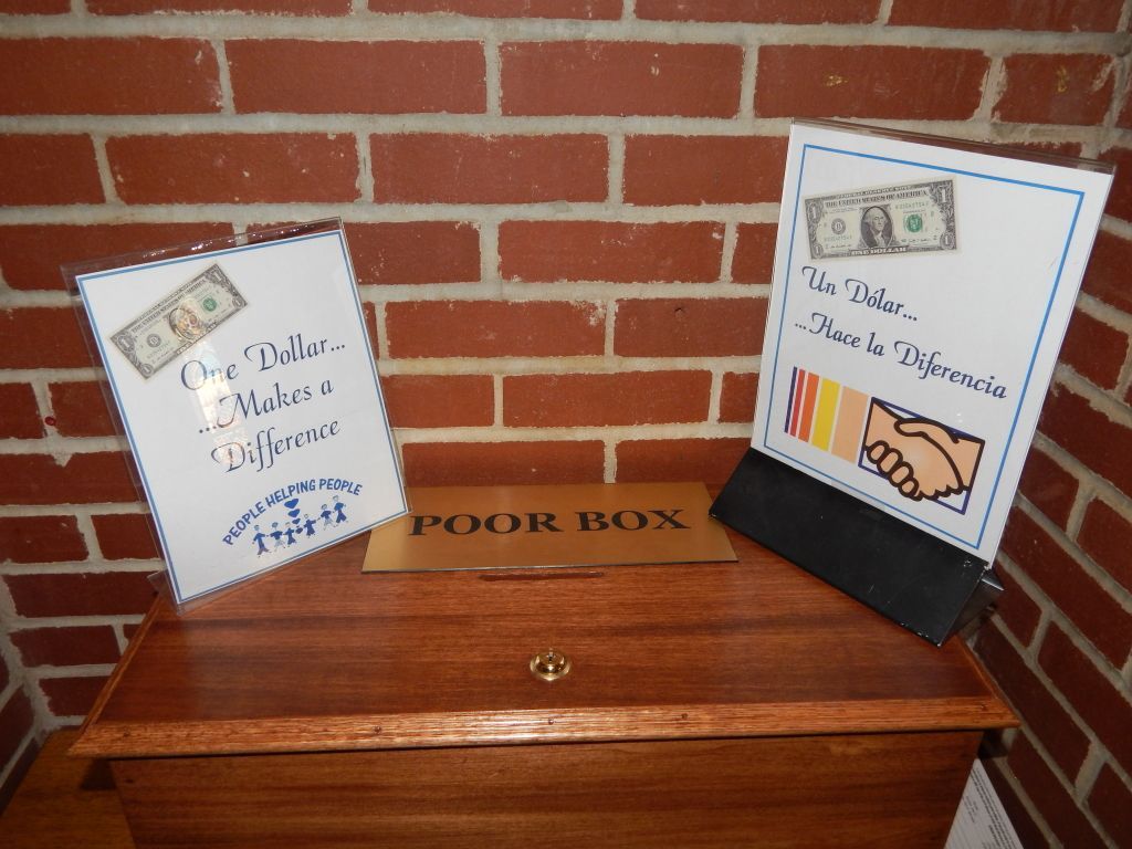 Wooden poor box with two donation signs against a brick wall, including cash and handshake illustrations