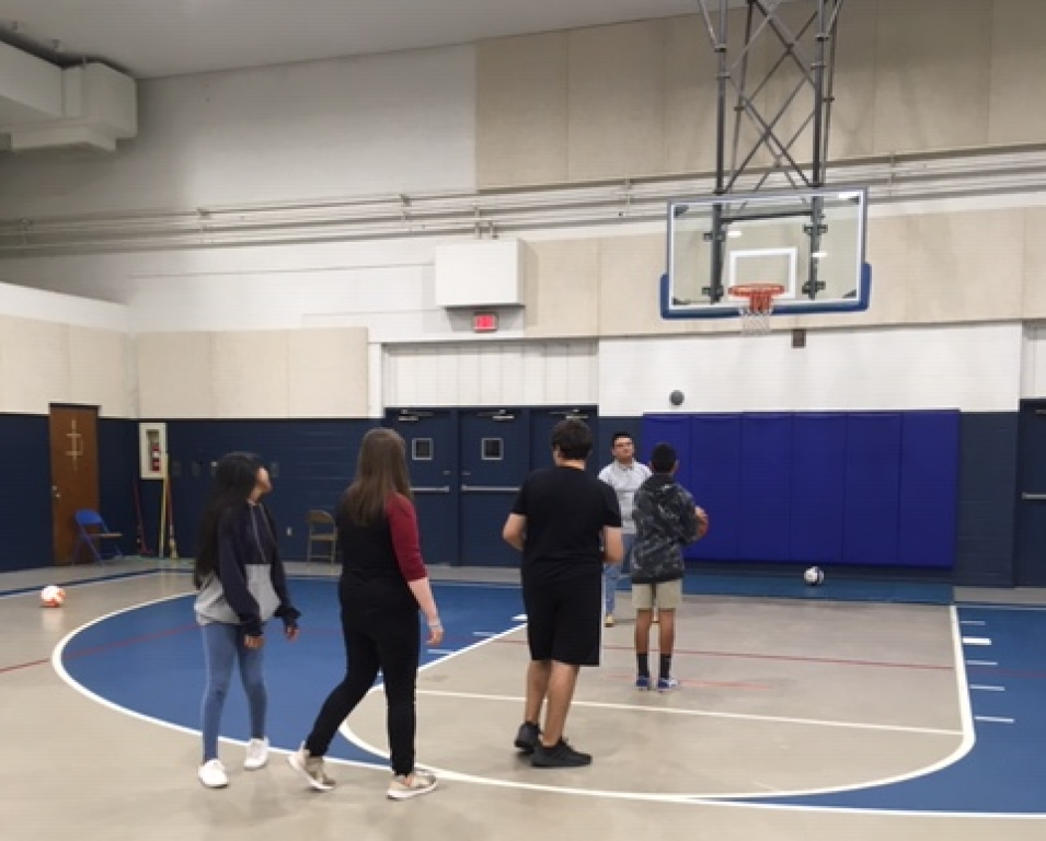 People standing on an indoor basketball court near the hoop, talking in a gymnasium