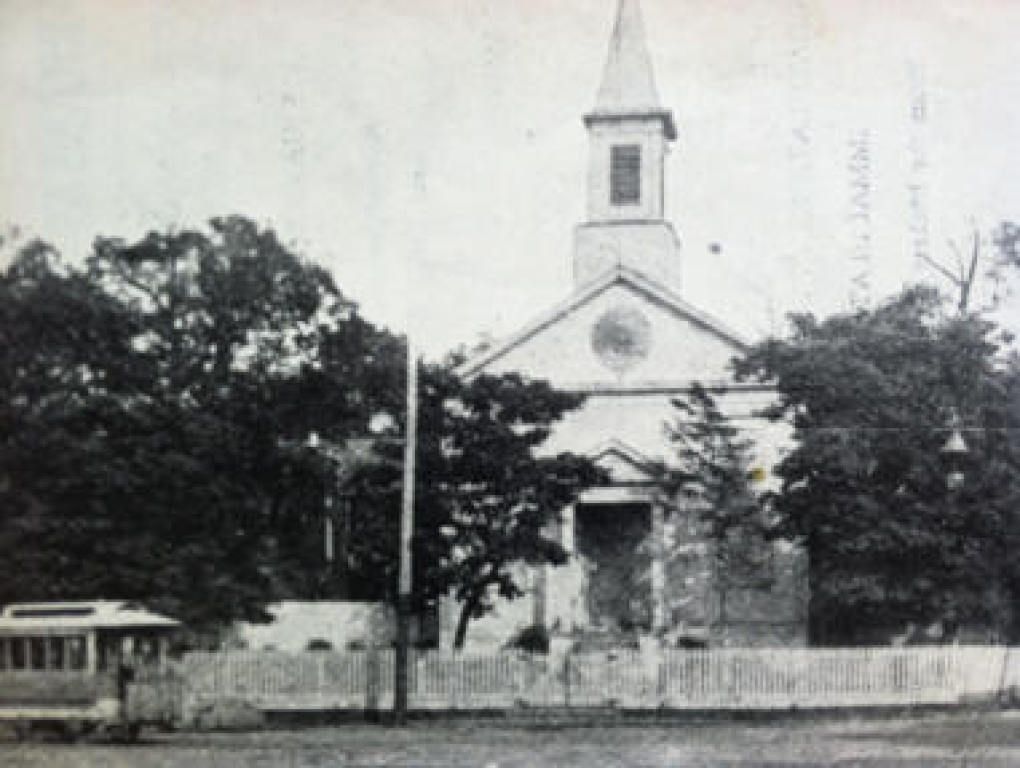 Black-and-white photo of a small church behind a white picket fence and trees.
