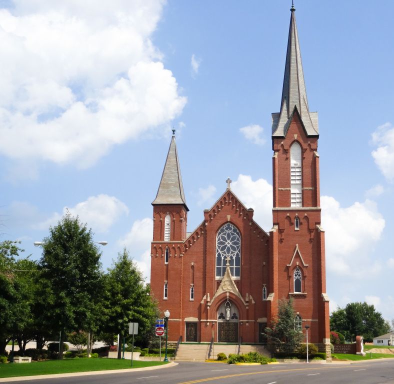 Red-brick church with tall steeple and arched windows on a sunny street corner