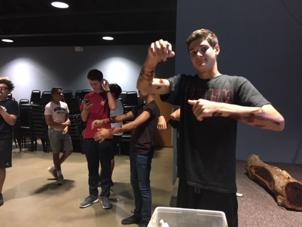 Group of people indoors; one man in a black T-shirt poses with raised hands in the foreground.