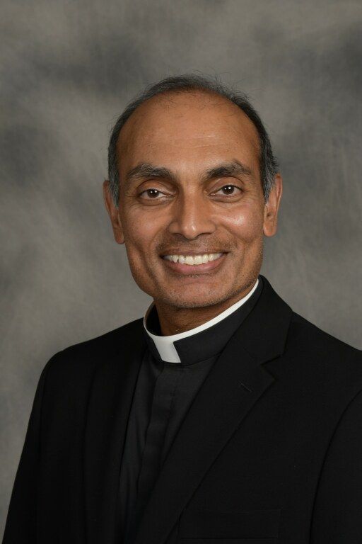 Portrait of a smiling clergyman in a black clerical suit and white collar against a gray background