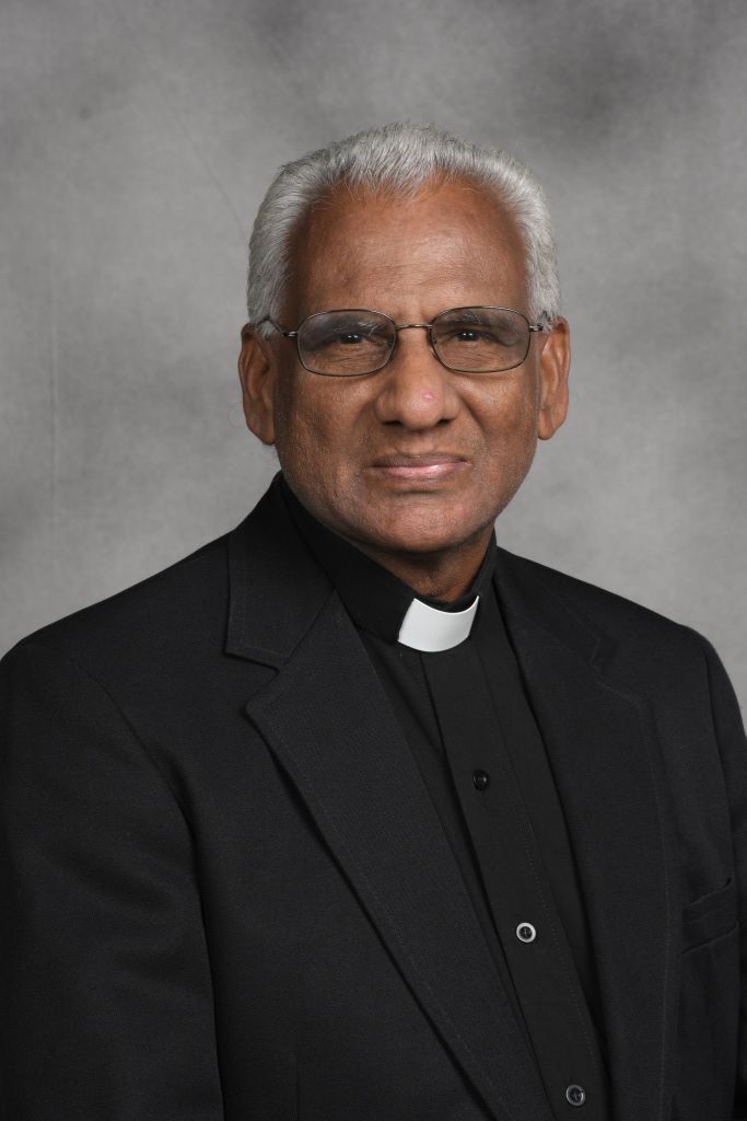 Portrait of a smiling clergyman in a black suit and clerical collar against a gray background