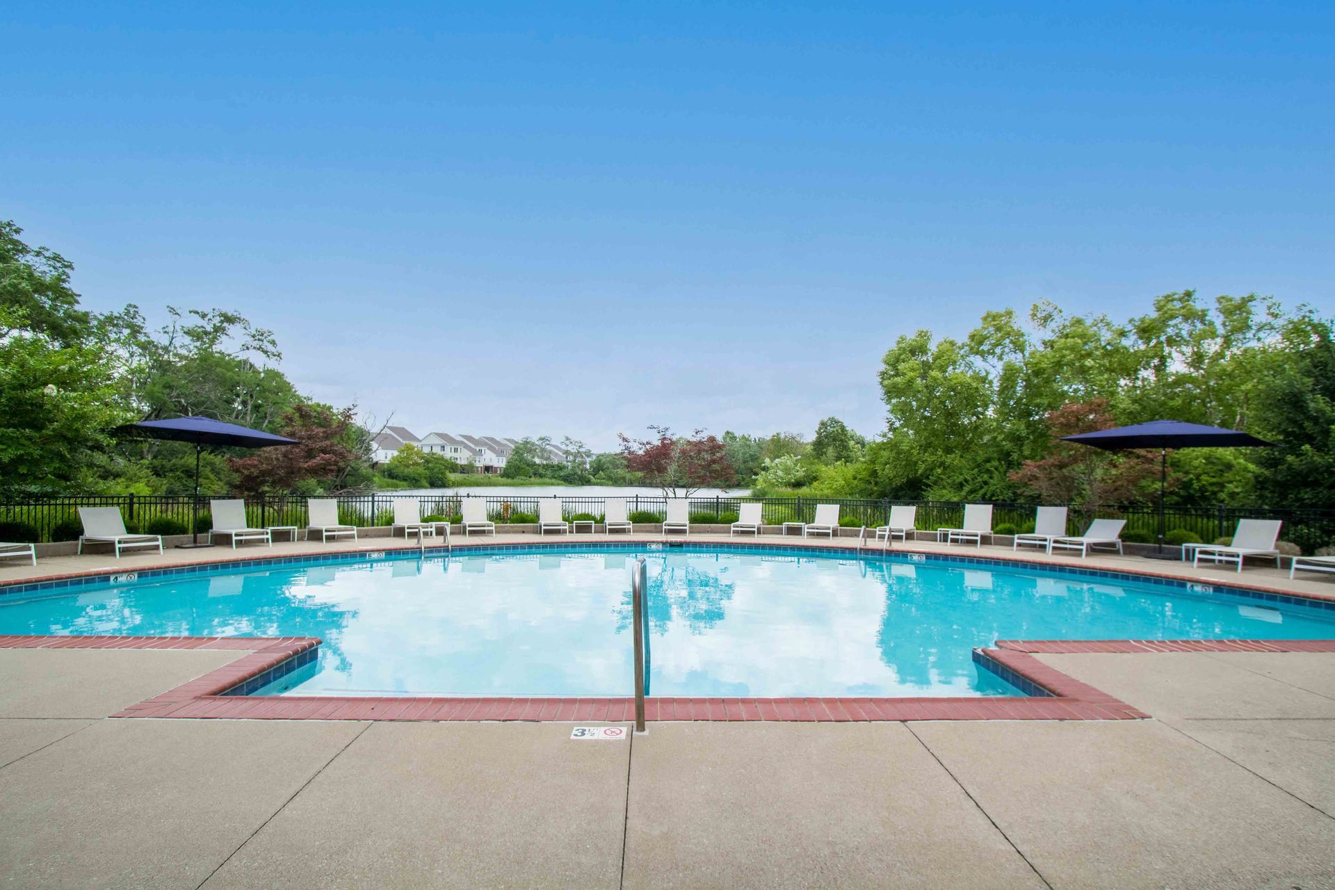 Swimming pool with lounge chairs under blue umbrellas, surrounded by trees under a clear sky at Hamburg Farms, offers apartments for rent in Lexington, KY.
