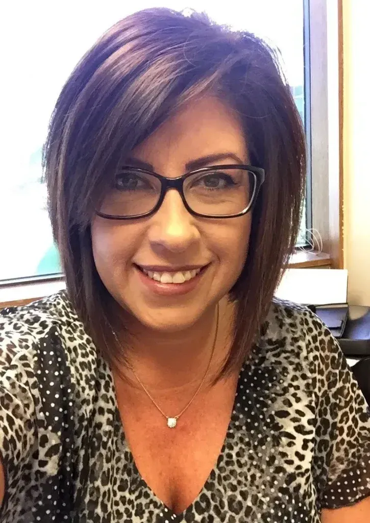 Woman with dark hair and glasses smiles; wearing patterned shirt, necklace.