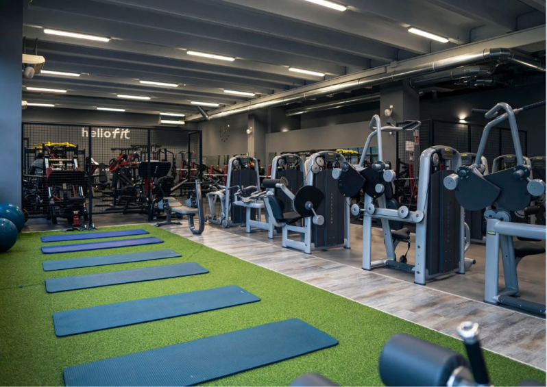 Interno di una palestra con attrezzi ginnici, pareti grigie e luci intense dall'alto.