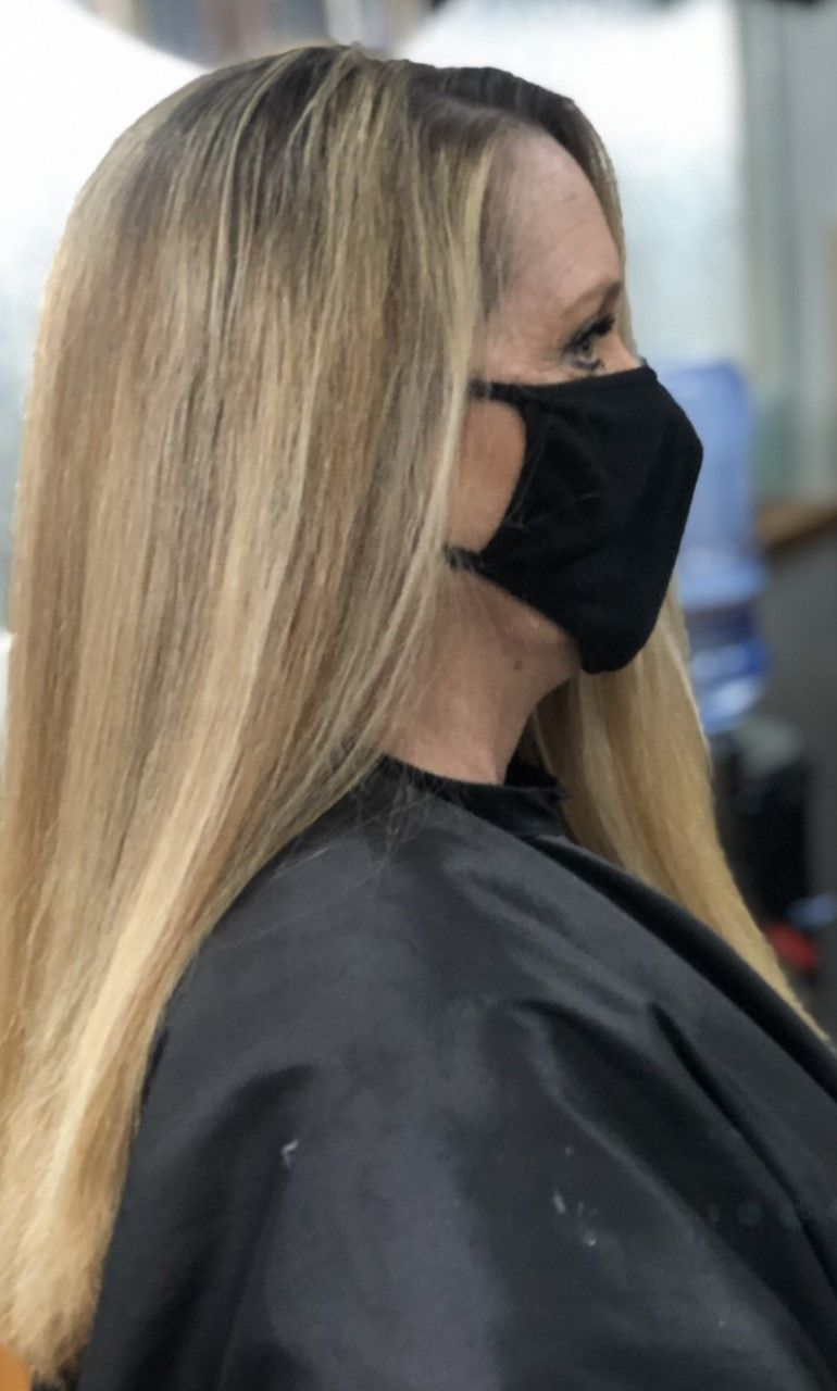 Woman with long, blonde hair wearing a black face mask and salon cape.