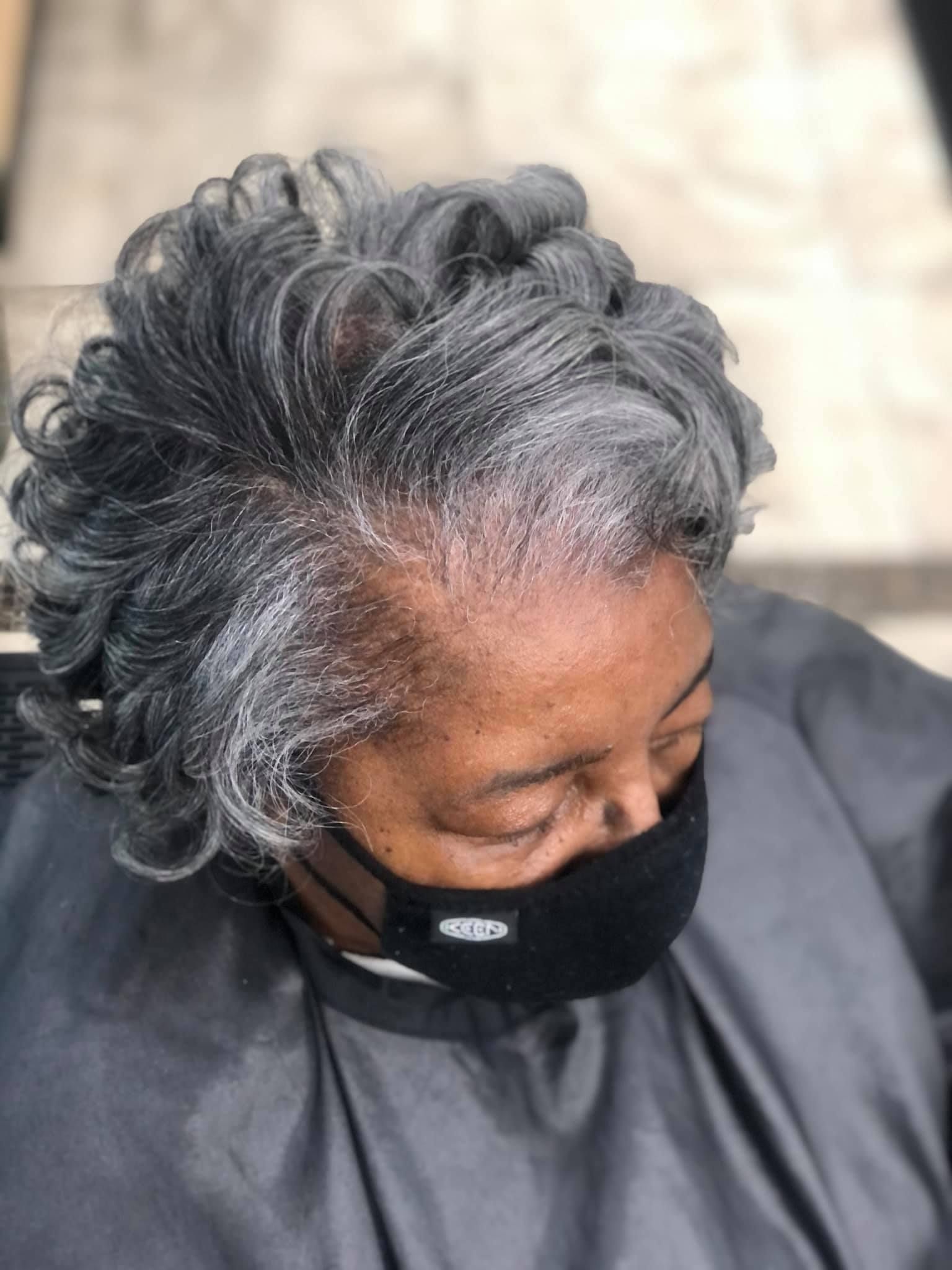 Person with short, wavy gray hair, wearing a black face mask, in a salon setting.