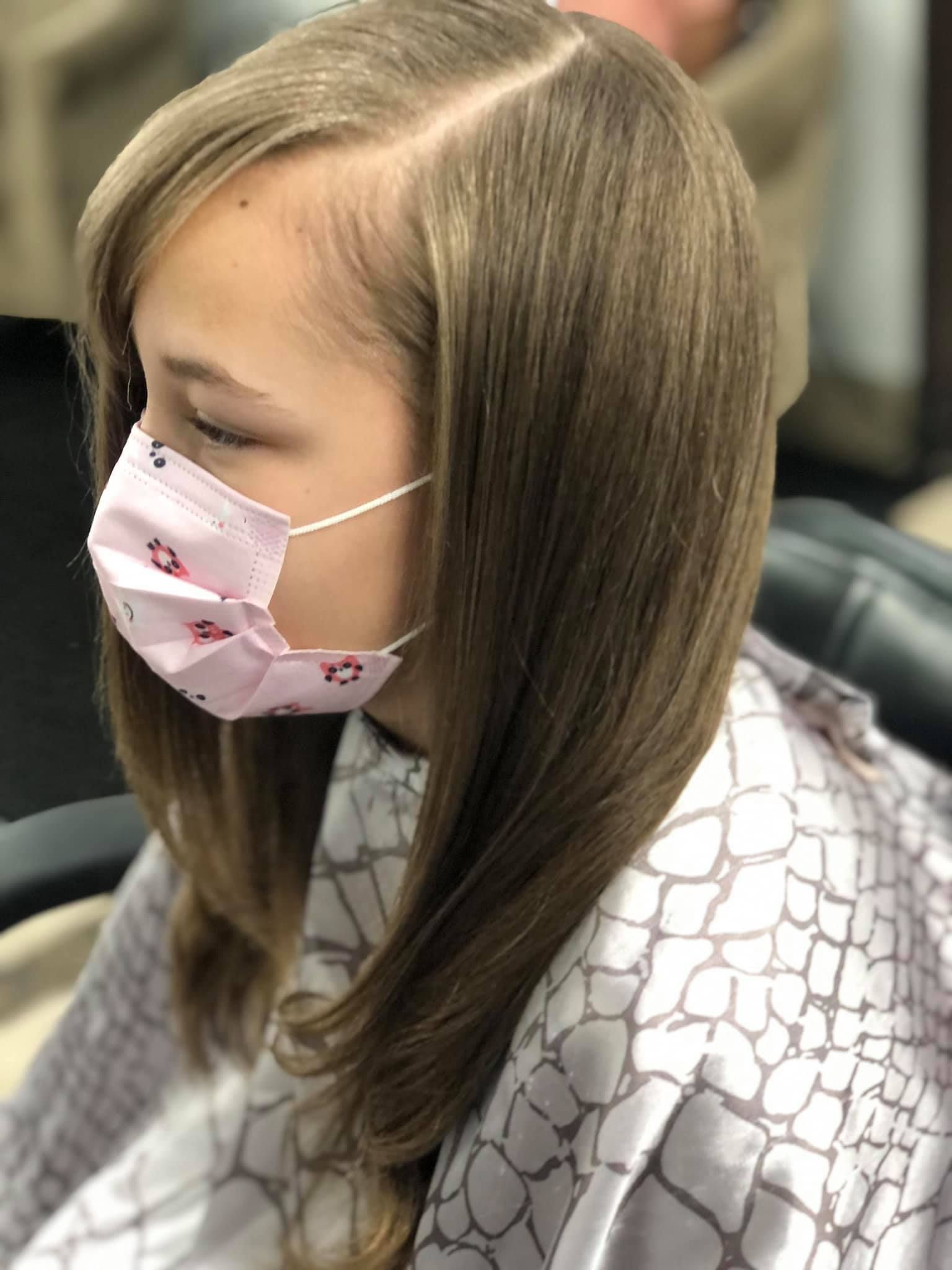 Girl with long, layered brown hair, wearing a pink face mask in a salon.