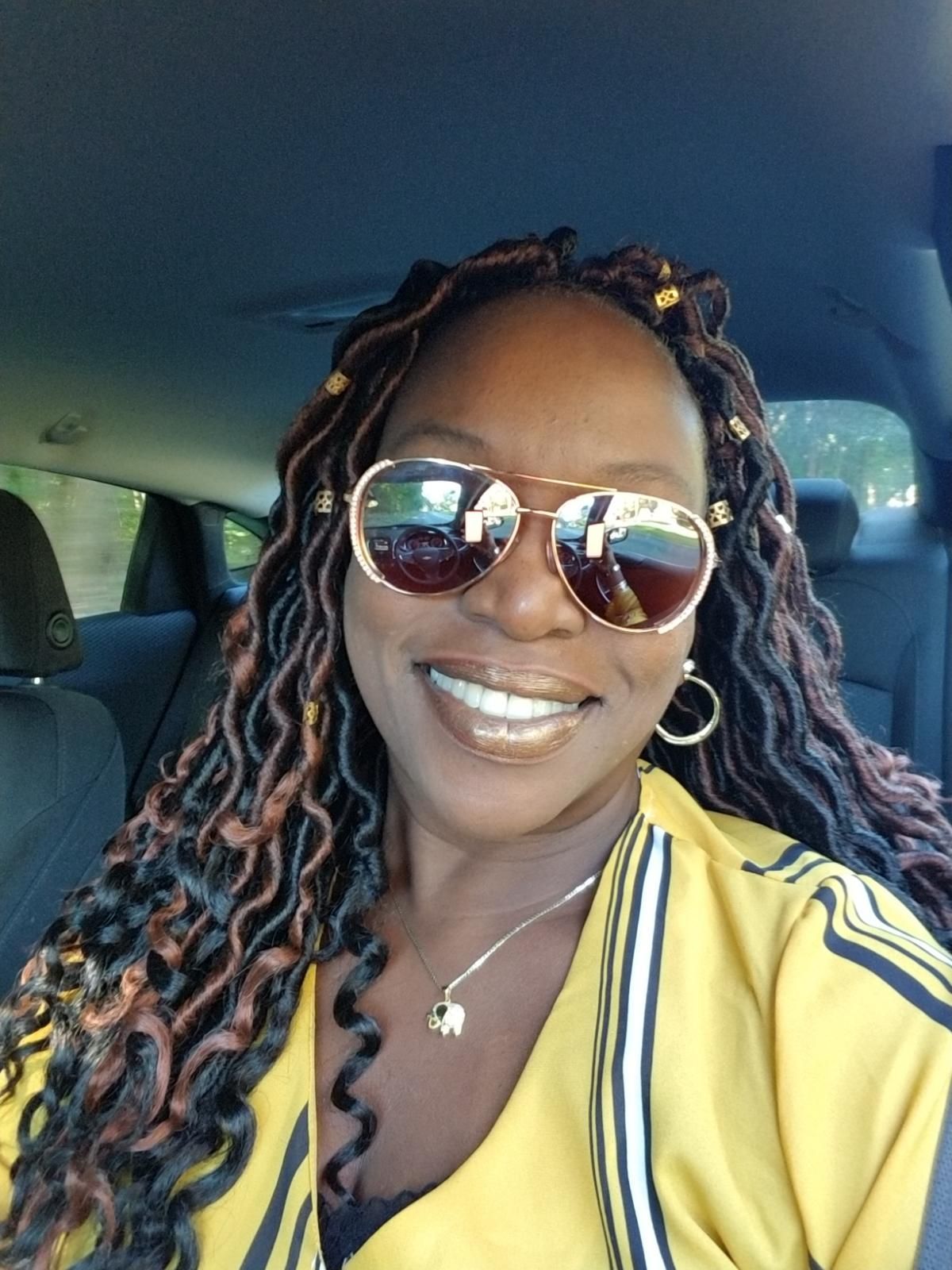 Person with locs wearing sunglasses, smiling, and wearing a yellow shirt, inside a car.