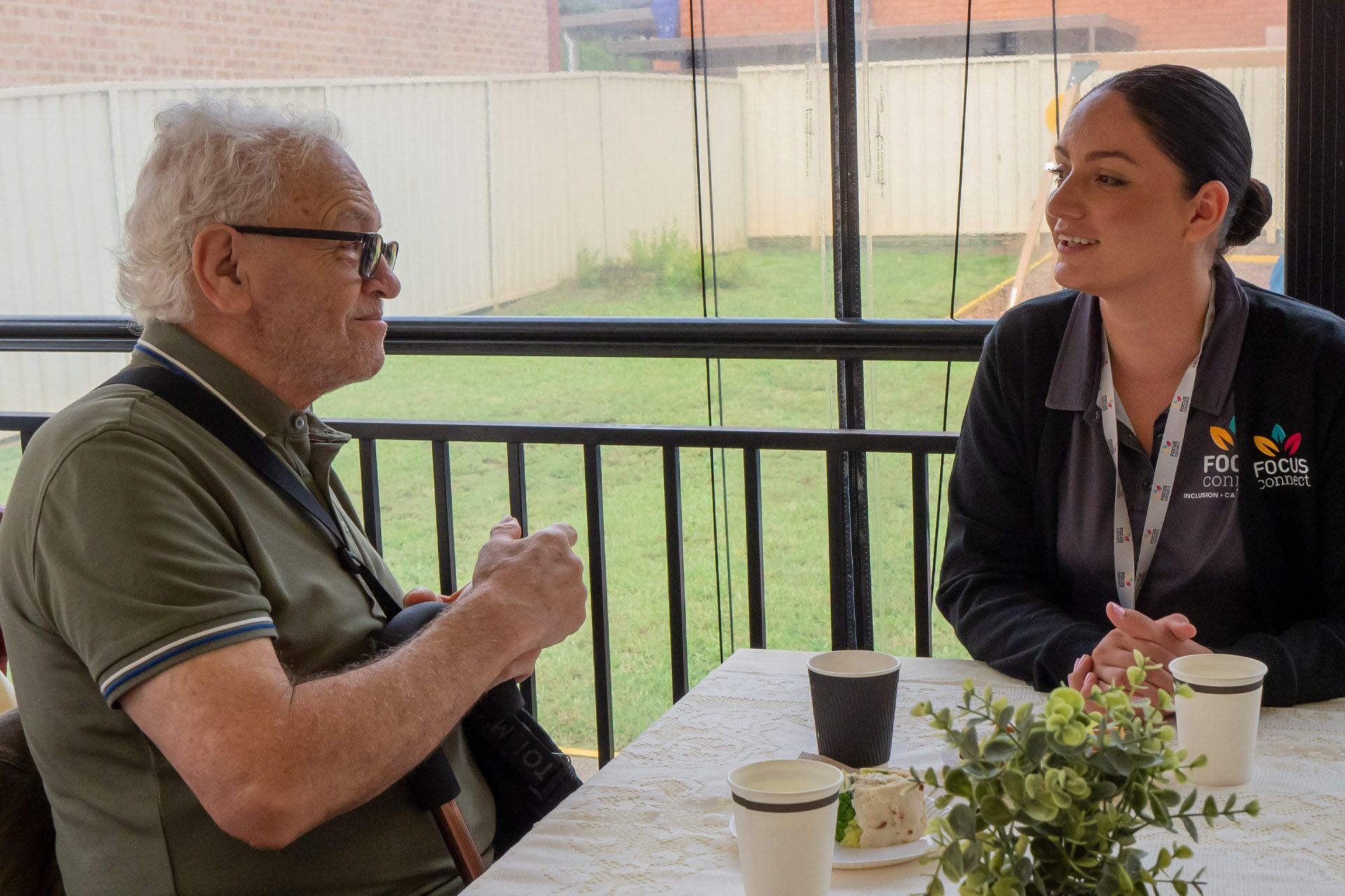 An older person and a staff member in a work uniform have a conversation at a table outside.