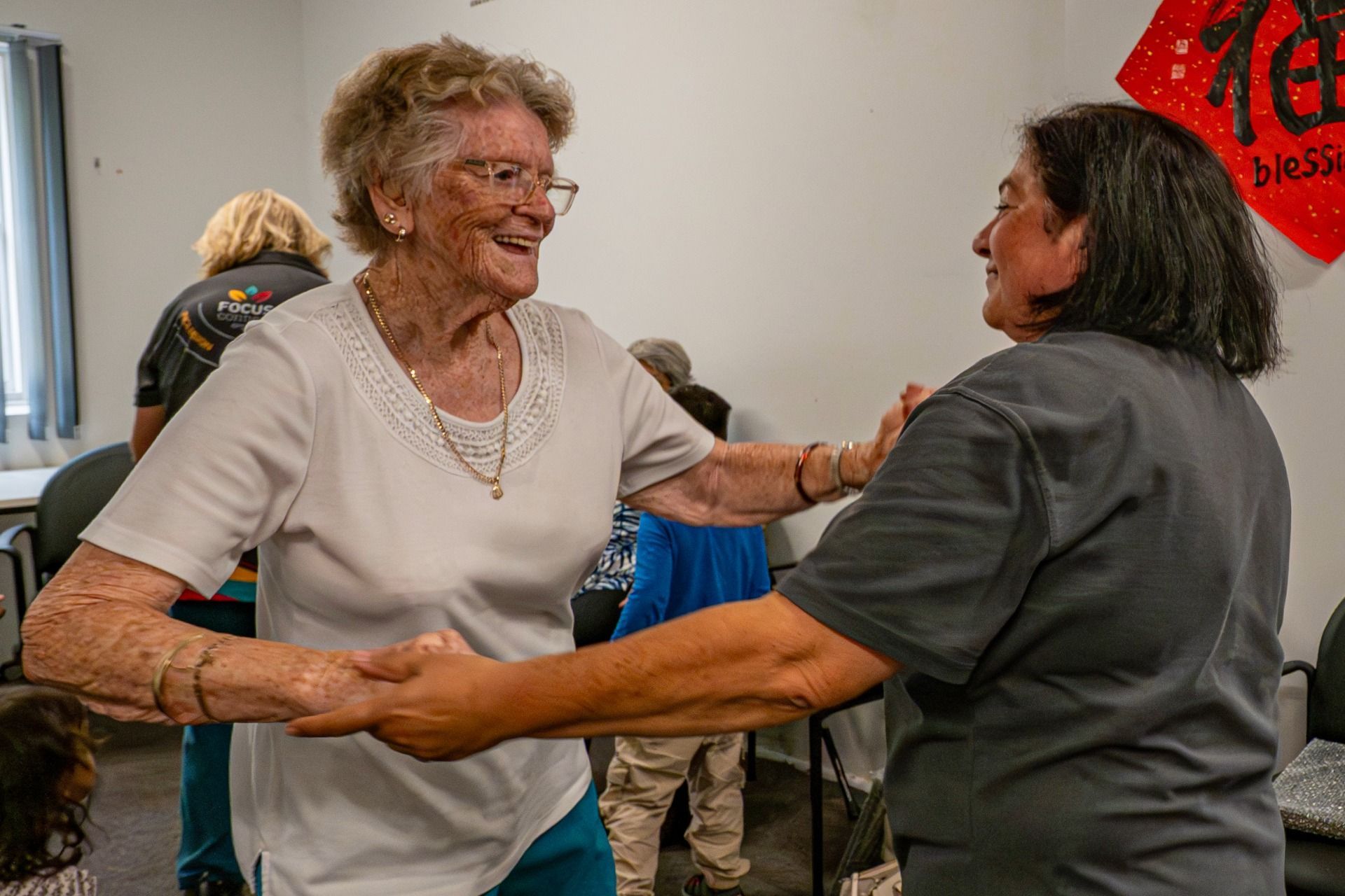 Two people hold hands and face each other in an indoor space with a red decoration on the wall, appearing to dance.