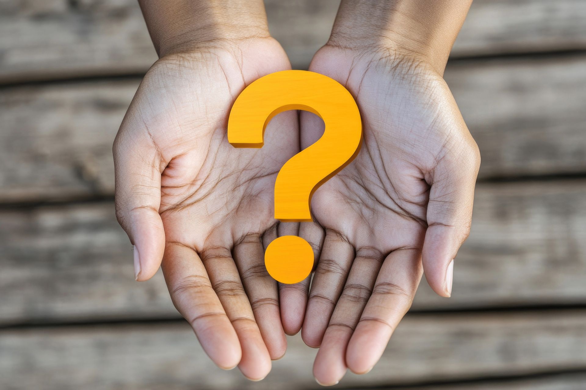 Hands cupping a bright yellow question mark, set against a blurred wooden background.