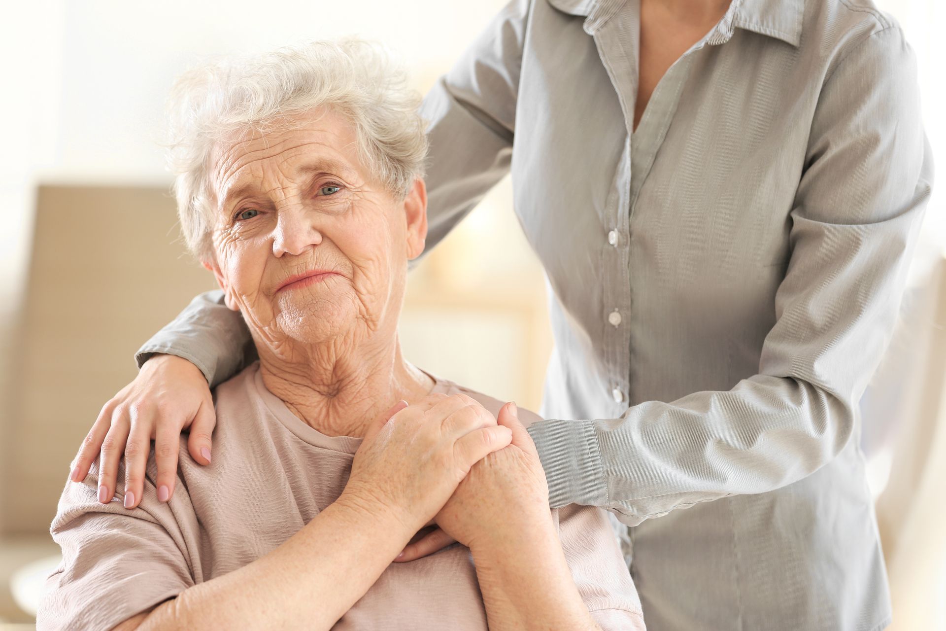 An elderly woman is sitting in a chair and a woman is putting her arm on her shoulder.