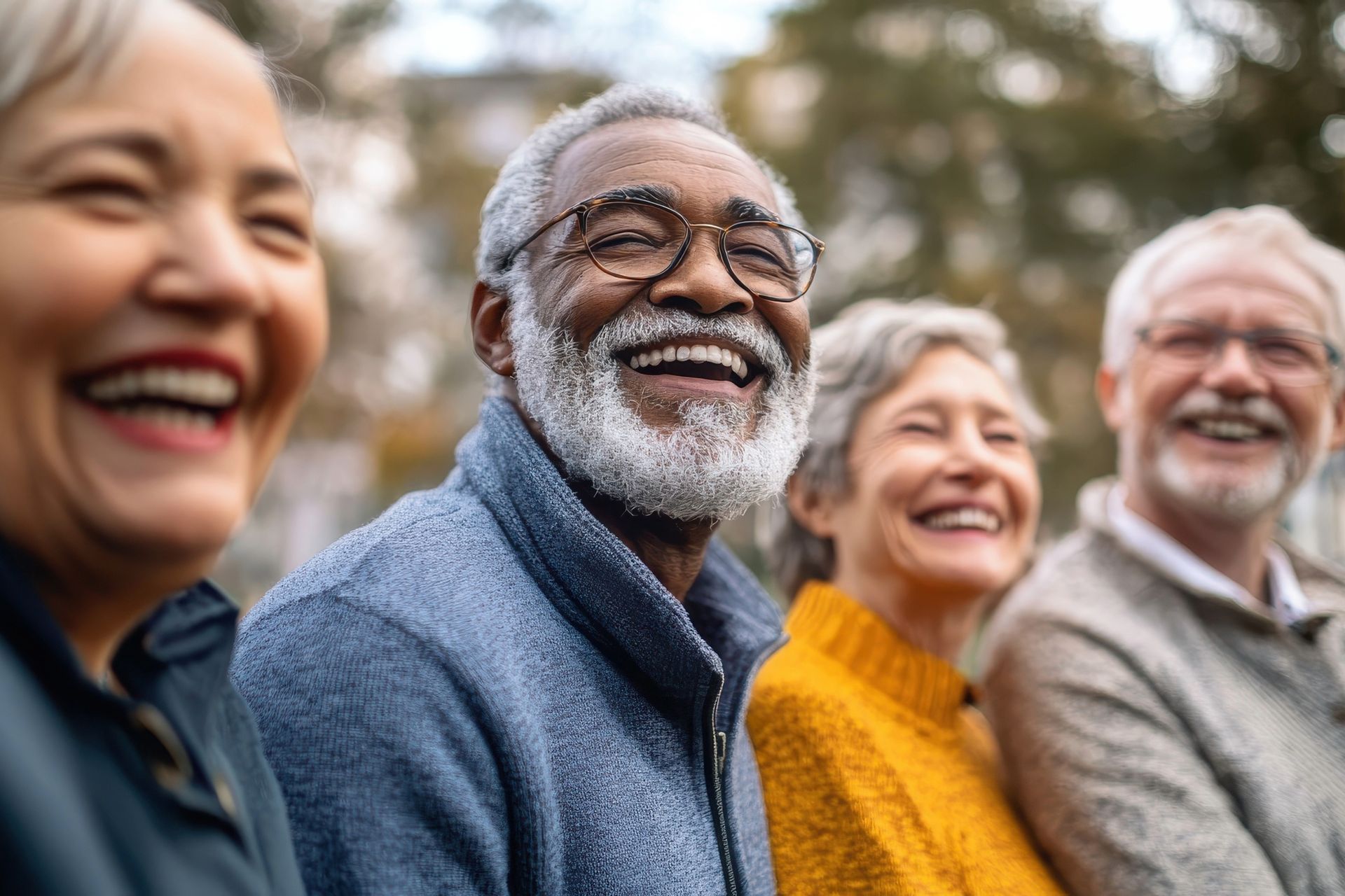 Four smiling older adults in an outdoor setting. They are diverse, with gray hair and visible joy.