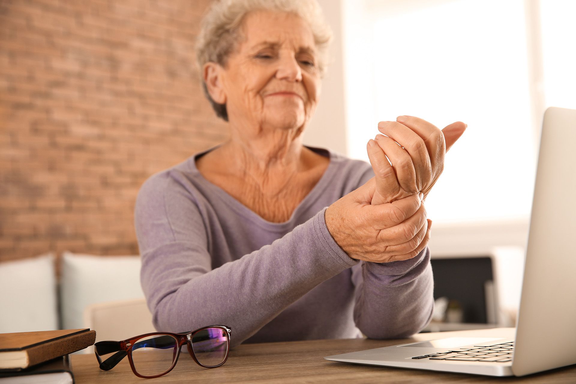 An elderly woman is sitting at a table with a laptop and holding her wrist.