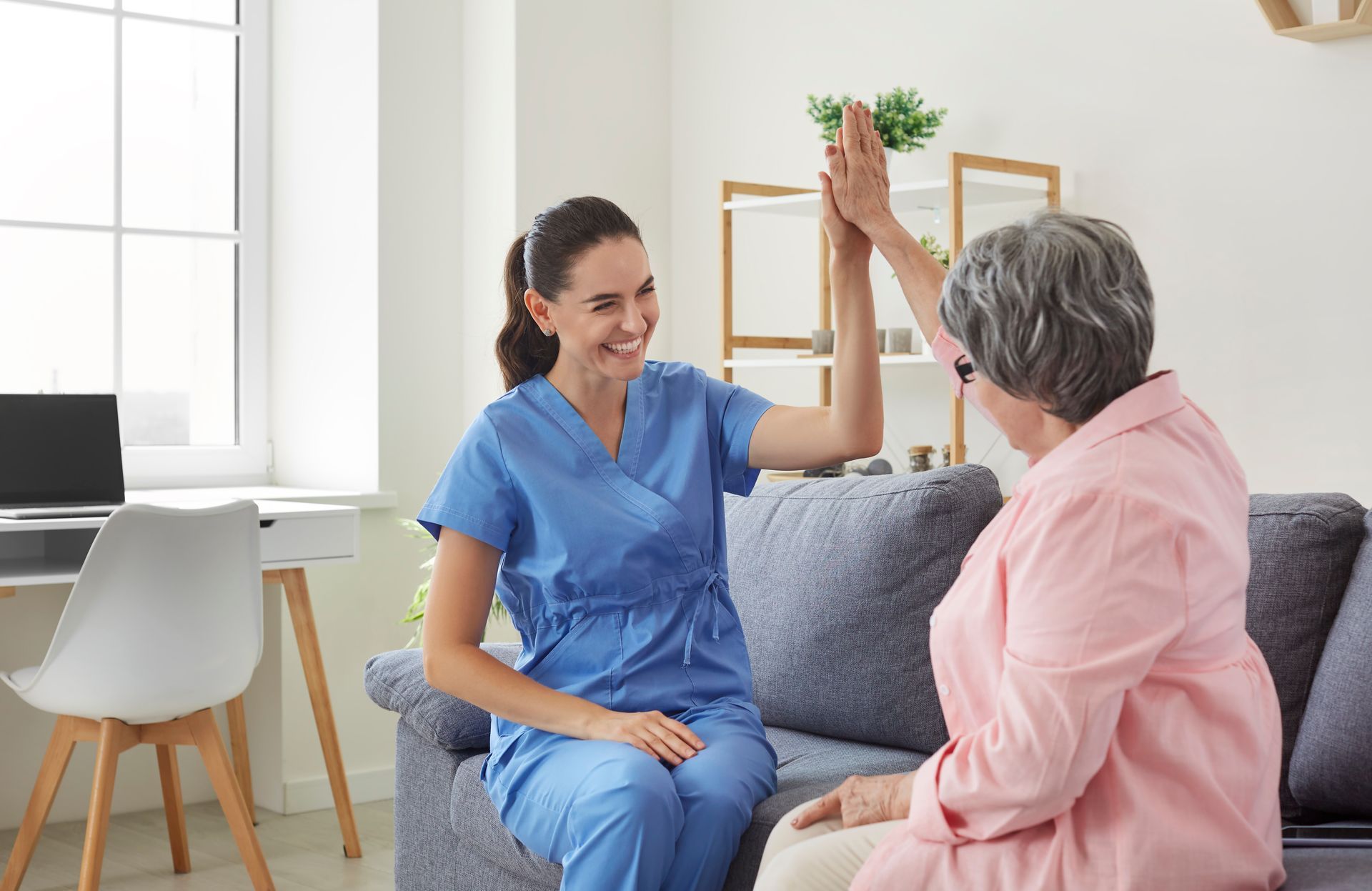 A nurse is giving an elderly woman a high five while sitting on a couch.