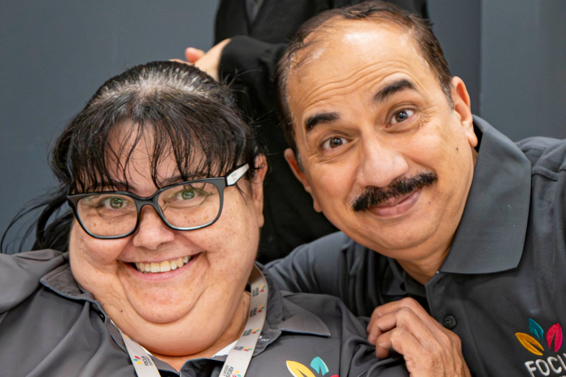 Two smiling people pose for a photo. They both wear gray polo shirts with a colorful logo. One person wears glasses.