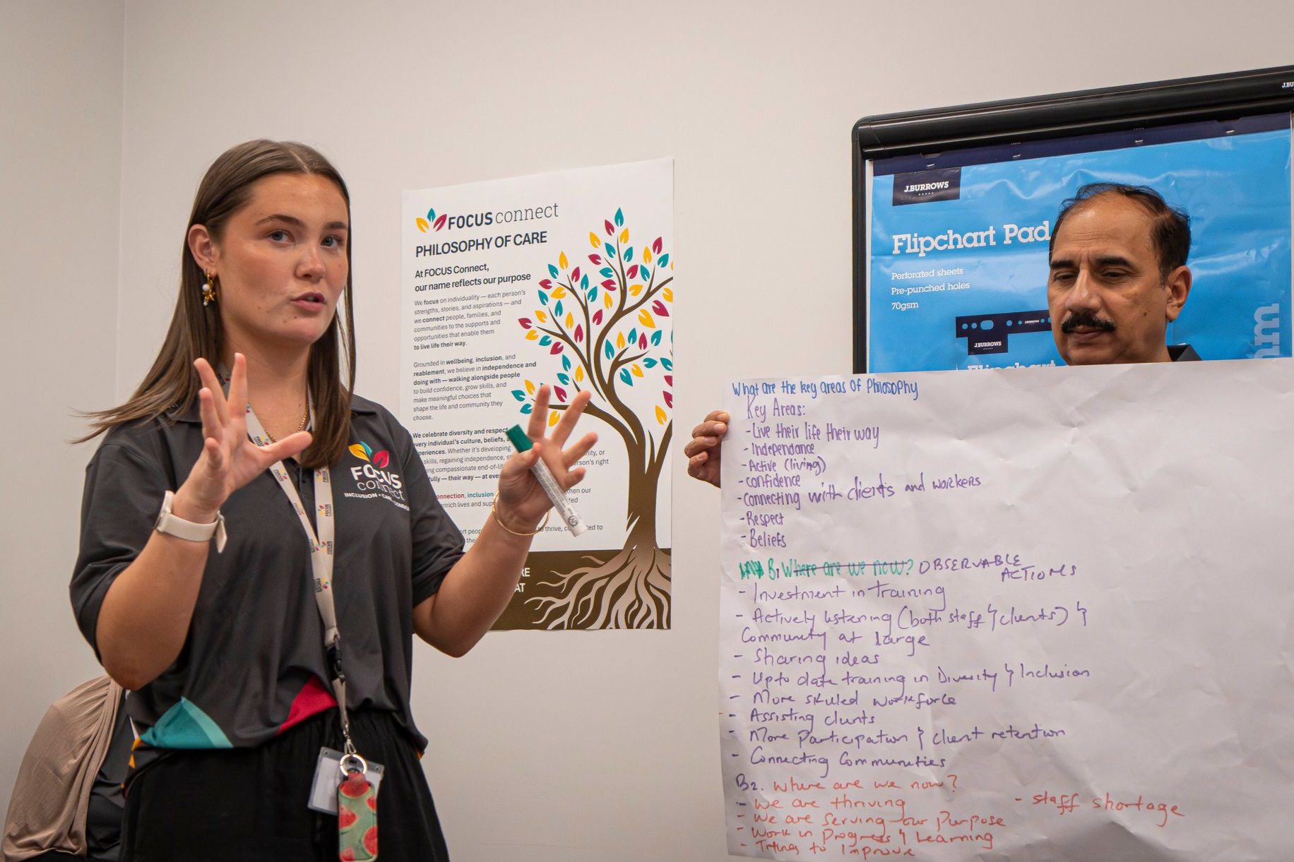 A person speaks while gesturing next to another person holding a whiteboard chart during an office presentation.