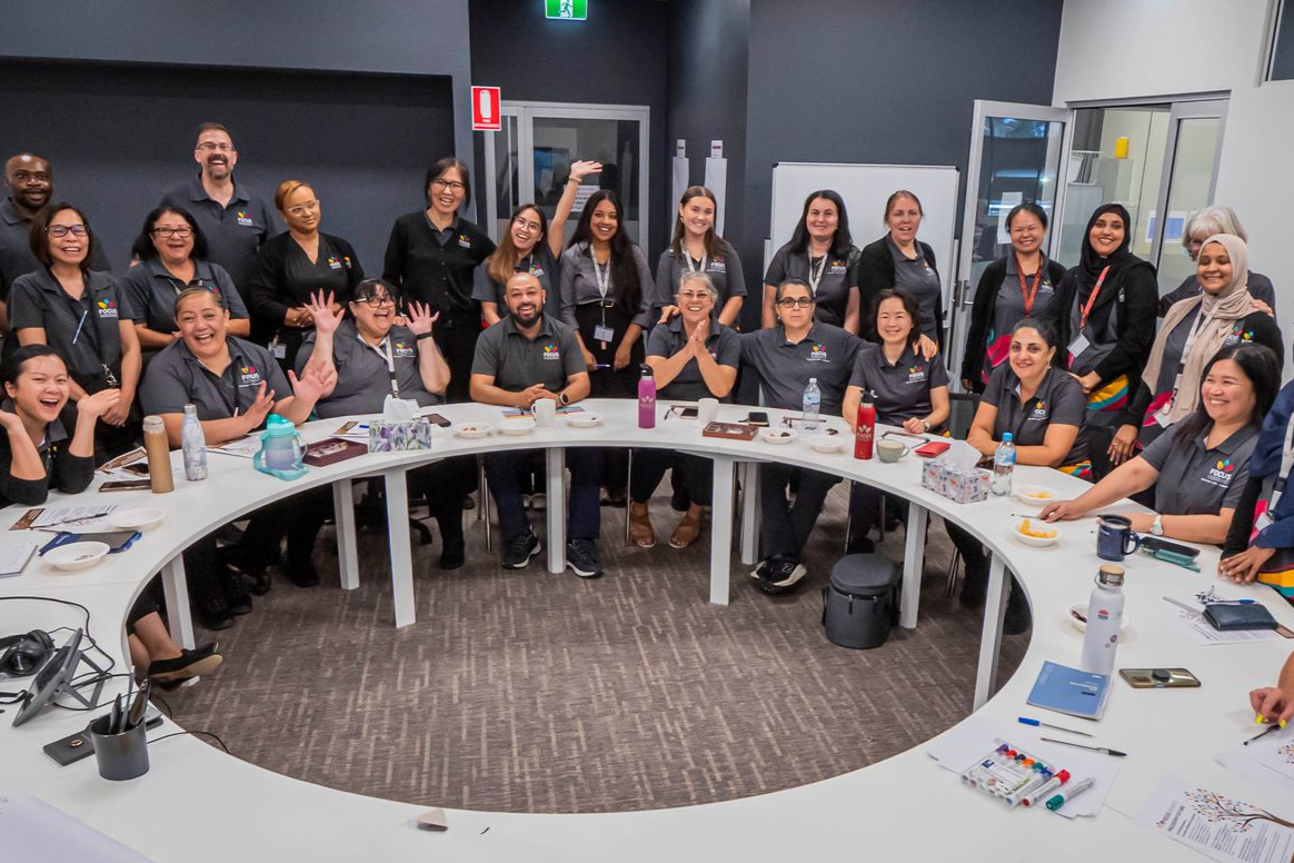 A large group of people smiling and posing around a semi-circular white table in a modern office meeting room.