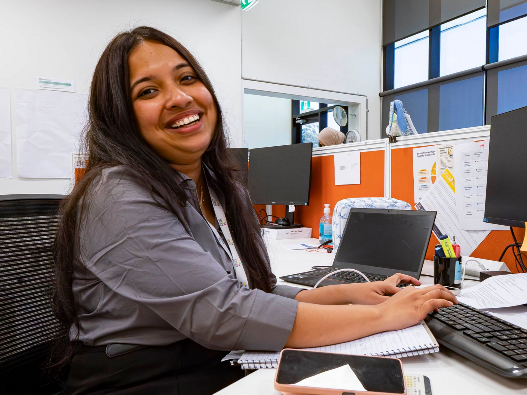 A smiling person sitting at an office desk, typing on a keyboard with a laptop and documents nearby.