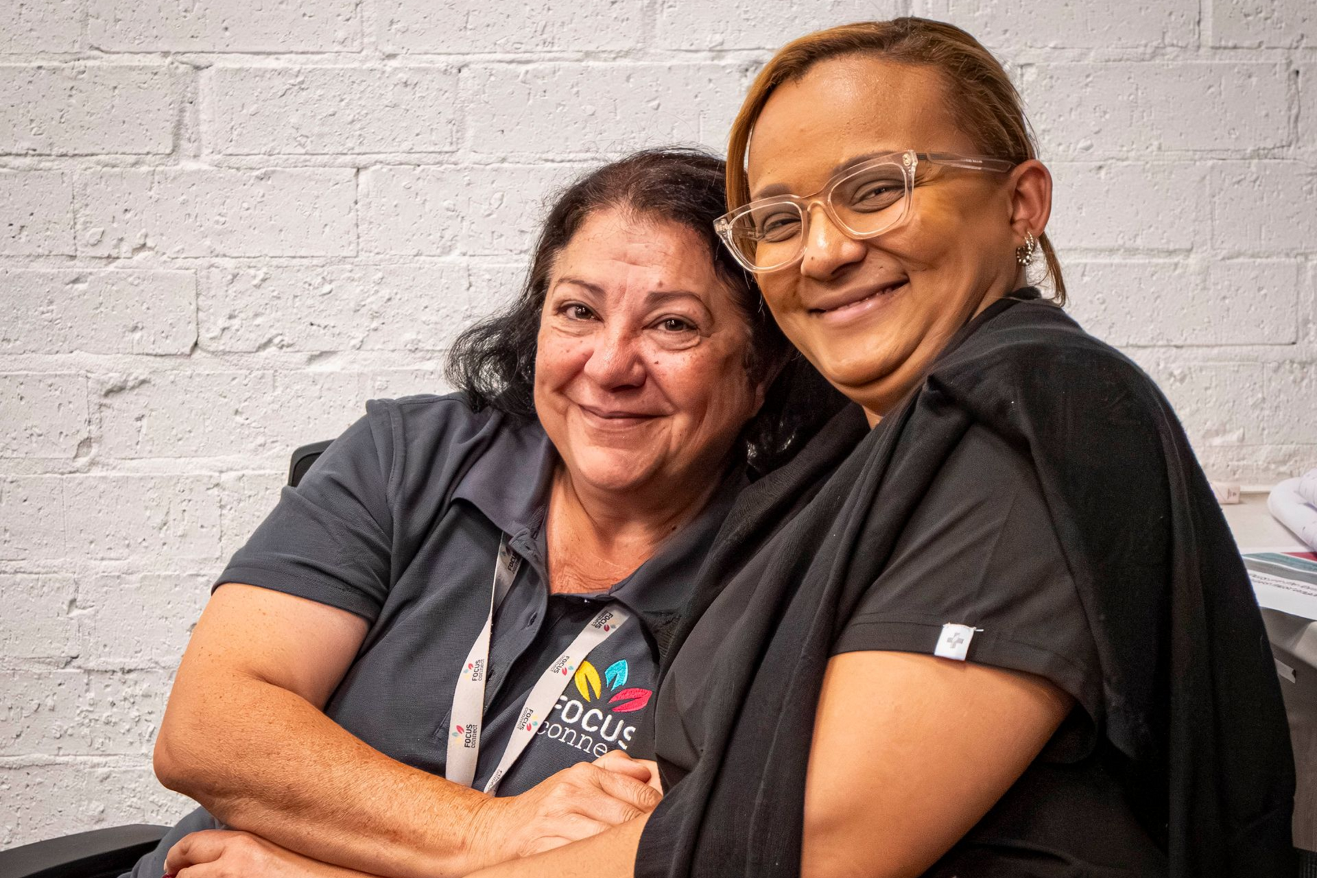Two smiling women embrace; one in a Focus Services shirt, the other in scrubs. White brick wall backdrop.