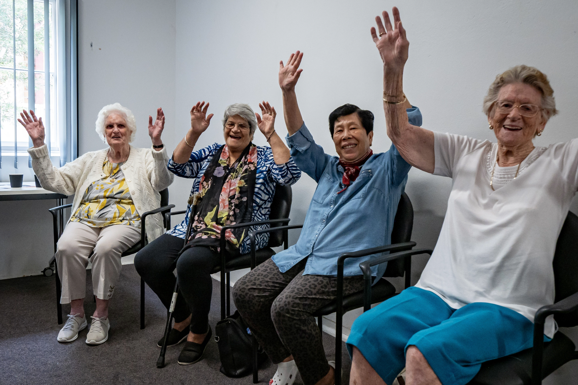 Five seniors seated indoors, smiling and raising their hands in a bright room