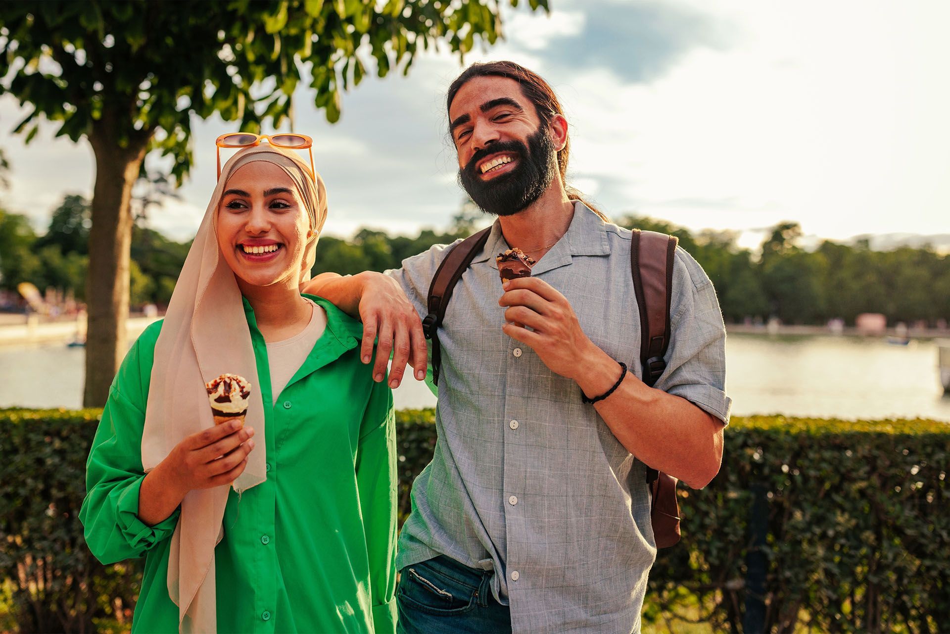 A man and a woman are standing next to each other in a park eating ice cream.