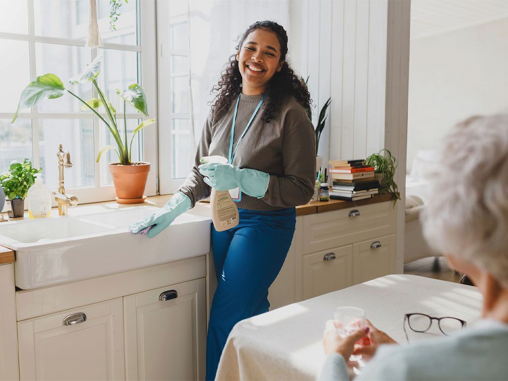 A woman is cleaning a kitchen counter next to an elderly woman sitting at a table.