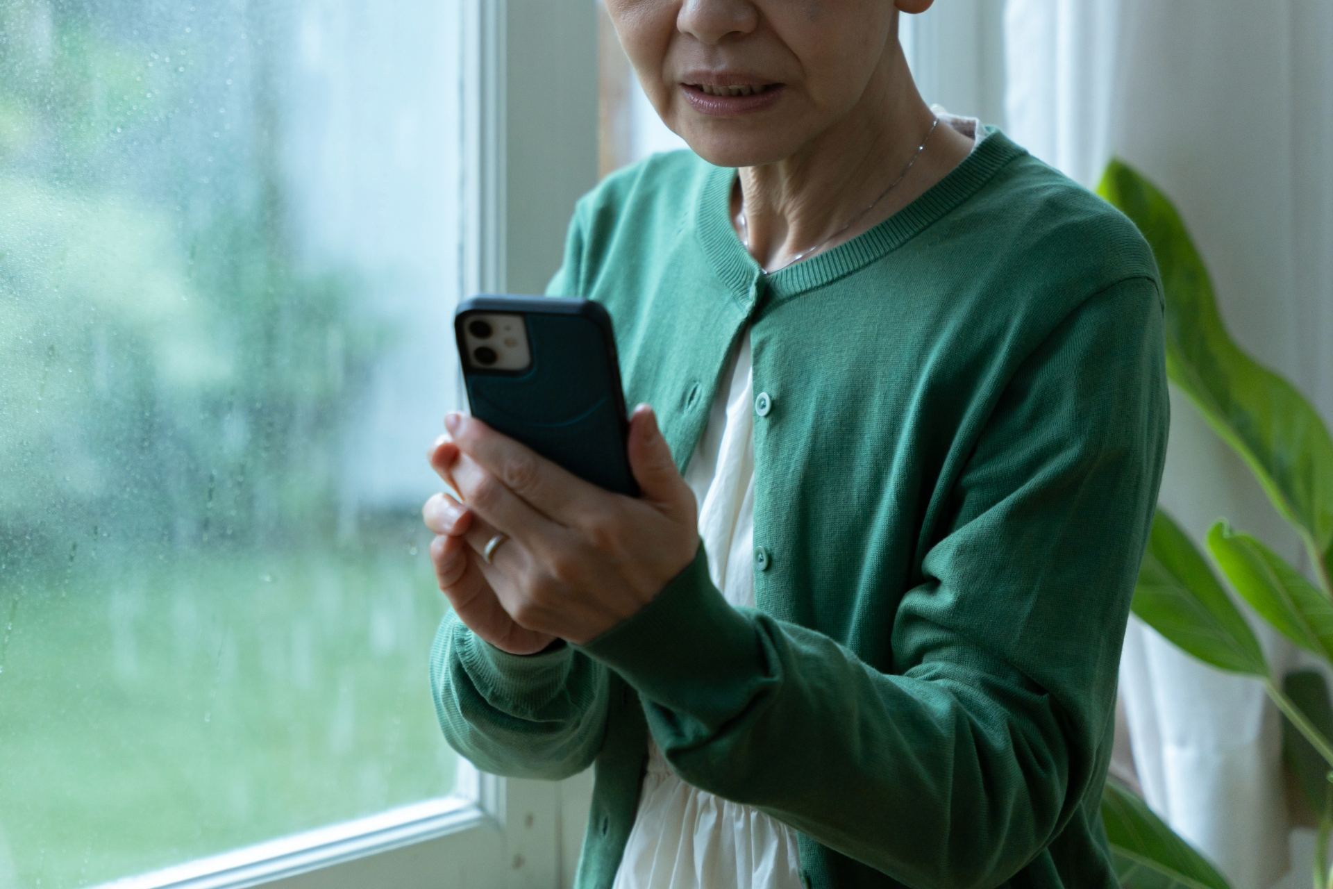 Senior in a green cardigan holding a smartphone by a window, looking at the screen.