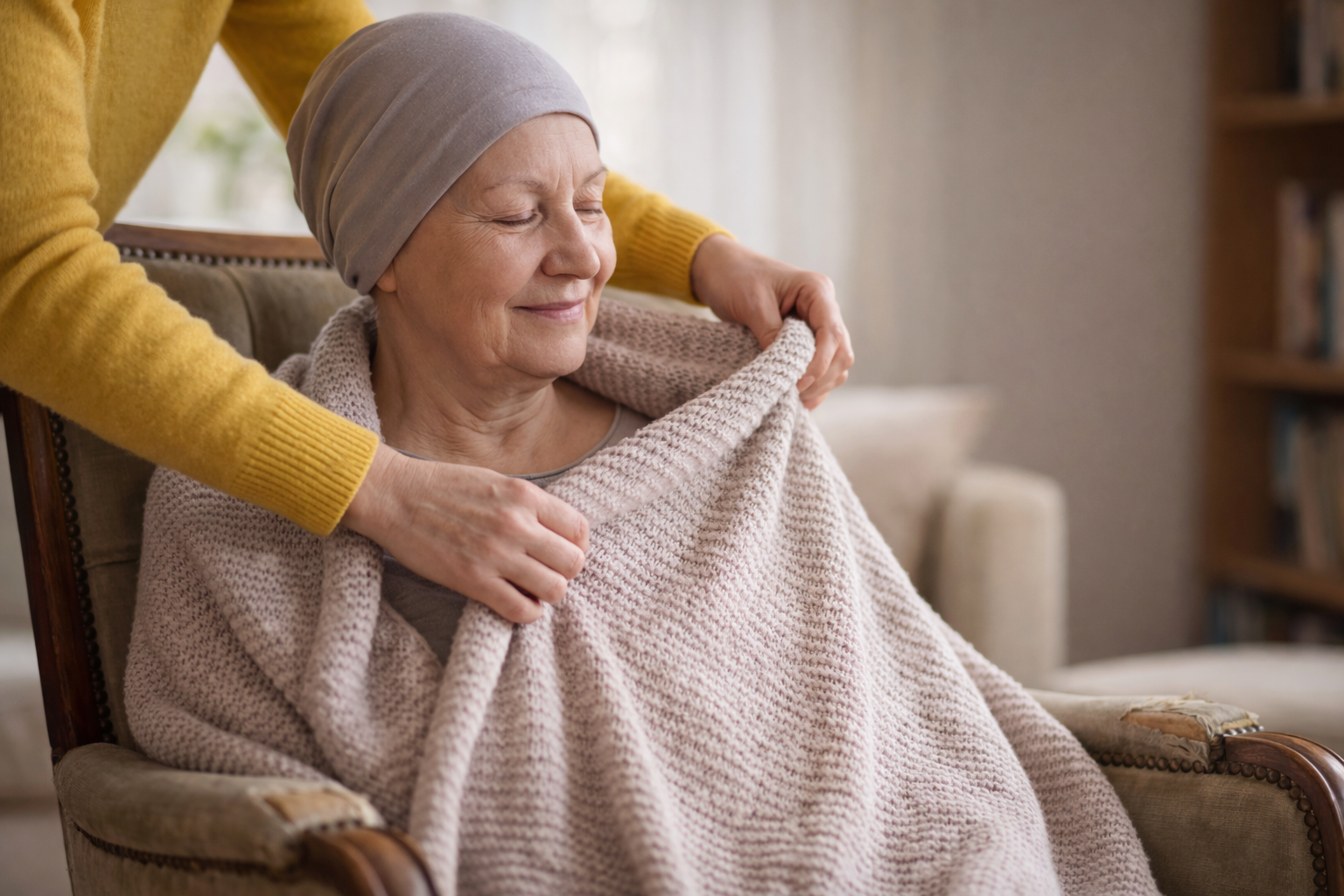Senior woman with head covering sits in a chair as someone covers her with a blanket.