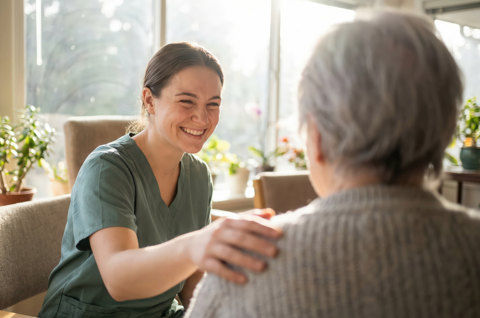 A caregiver in green scrubs smiling and placing a comforting hand on the shoulder of an older person in a bright room.