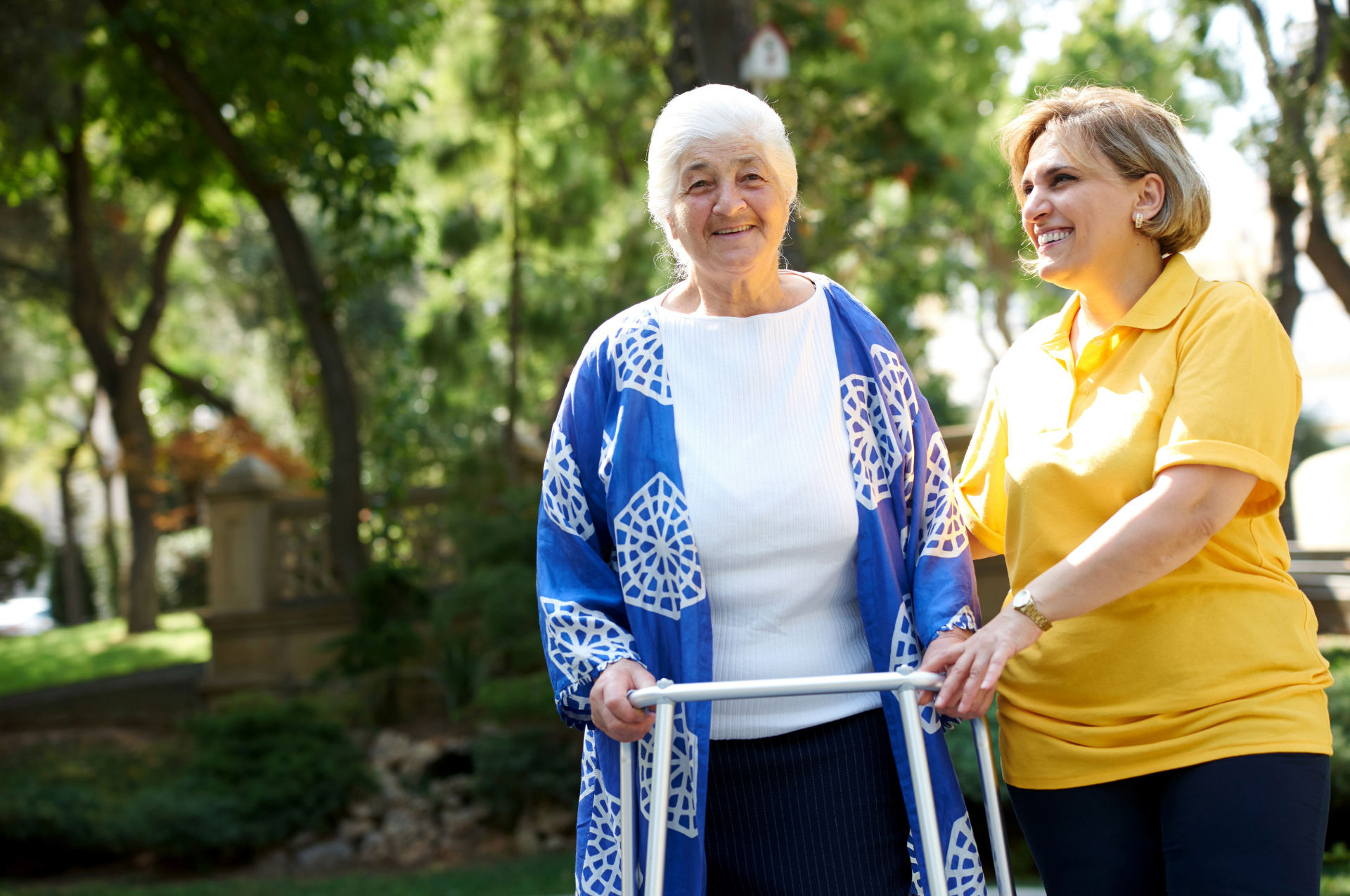 Woman with walker smiles outdoors with a caregiver in a yellow shirt.