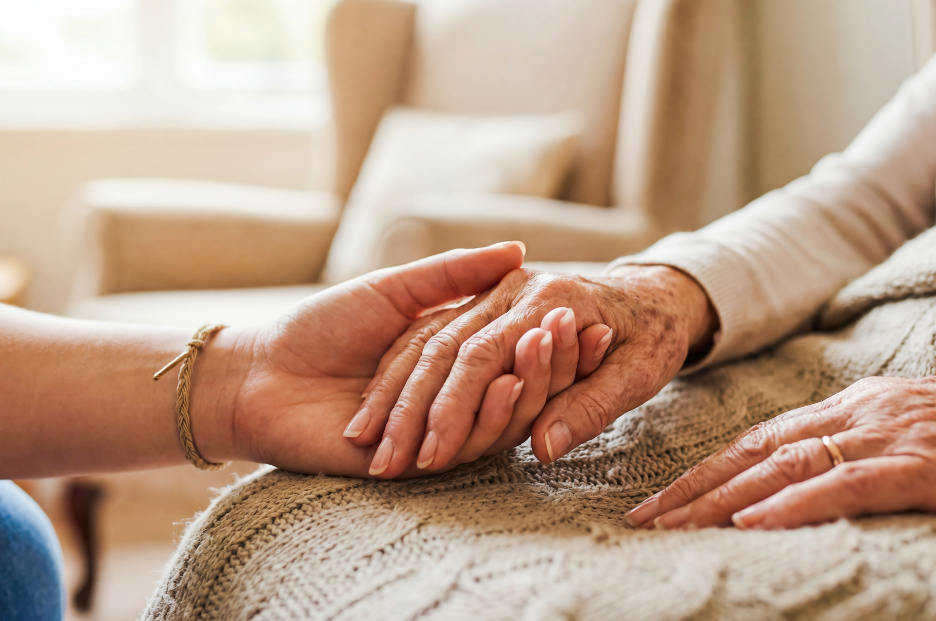 A hand holding an elderly person's hand, offering support. Indoors, warm lighting.