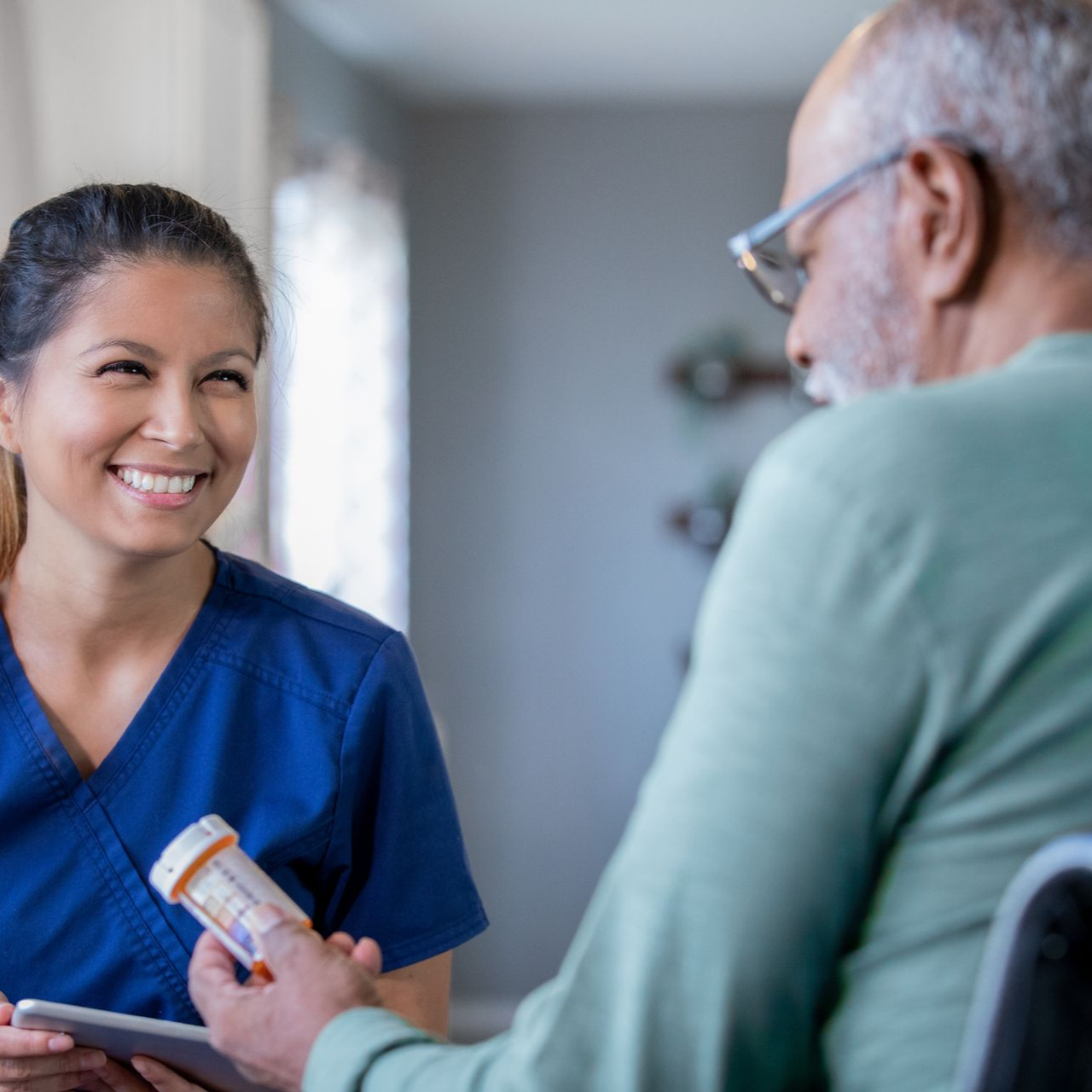 Woman in blue scrubs smiles at a man, handing him a pill bottle.