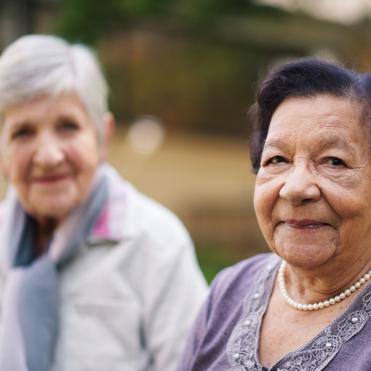 Two elderly women smiling outdoors; one in focus wearing purple, the other blurred.