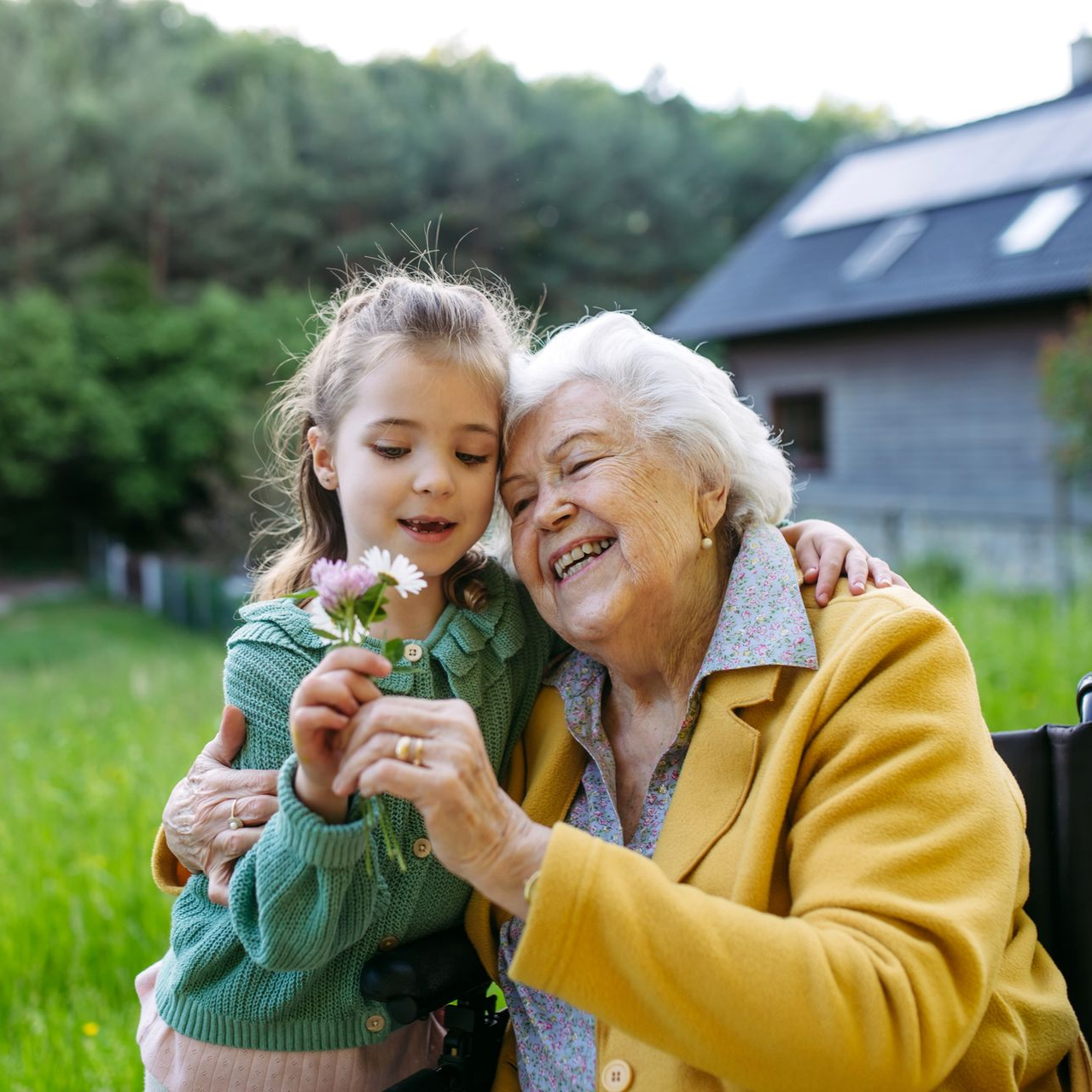 Girl hugs smiling elderly woman, holding flowers outside a house with green grass.