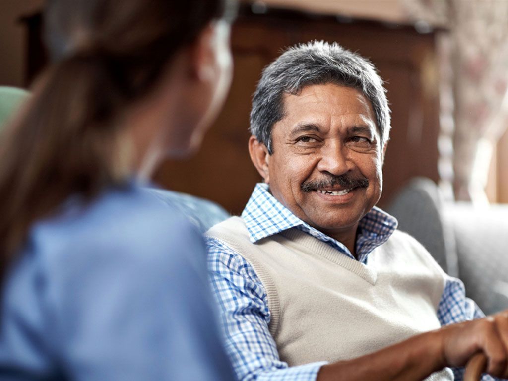 An elderly man is sitting on a couch talking to a nurse.