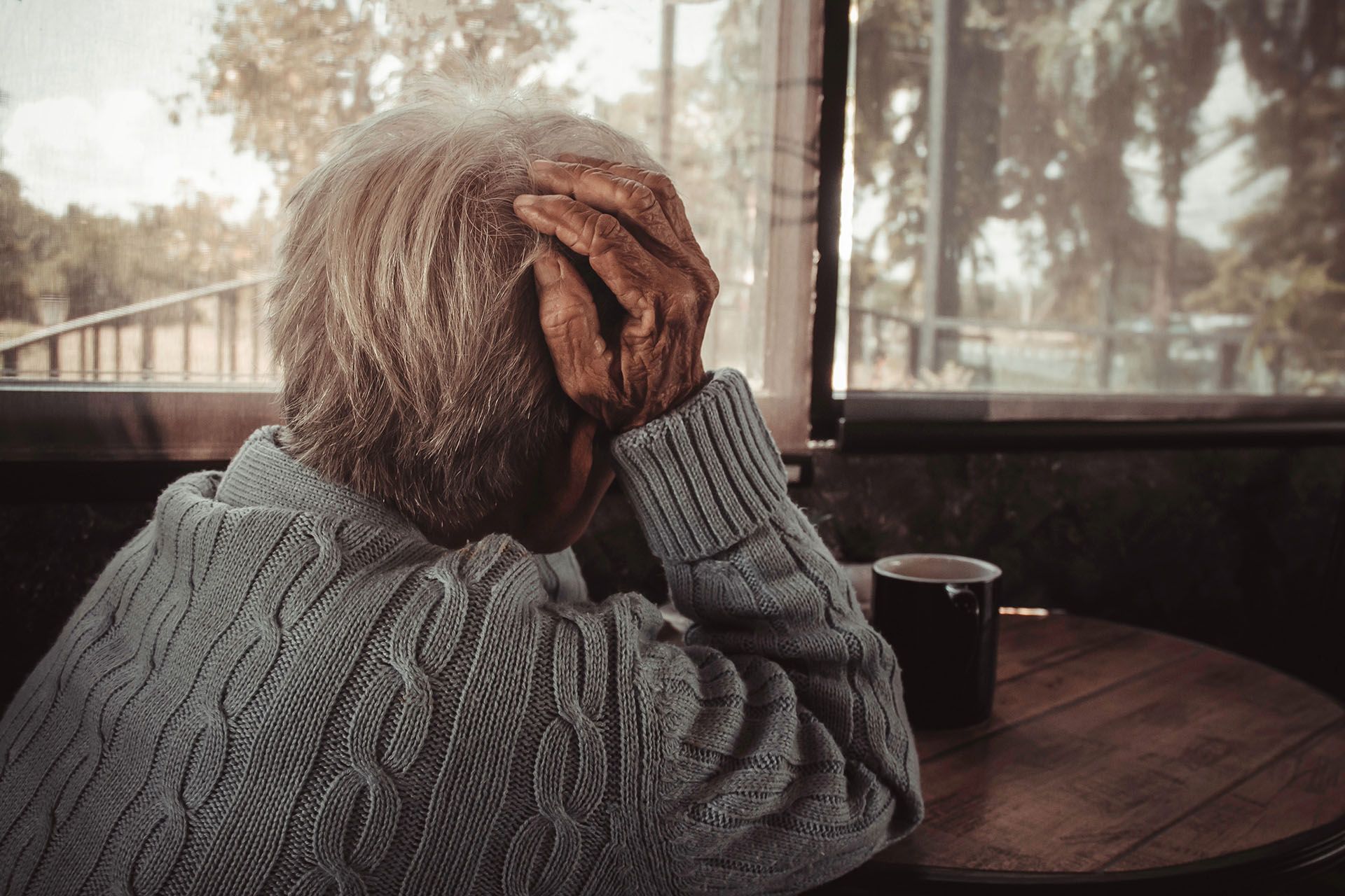 An elderly man is sitting at a table with his hands on his head.