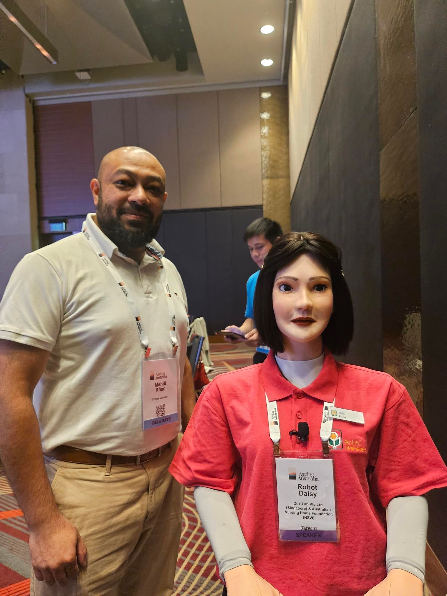 A man standing beside Robot Daisy, a humanoid wellness facilitator, at an aged care technology conference.