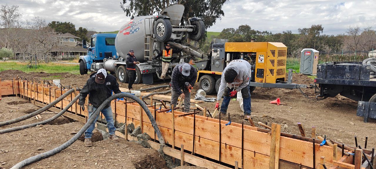 Construction workers pouring concrete beside a concrete truck at a foundation site with wooden forms