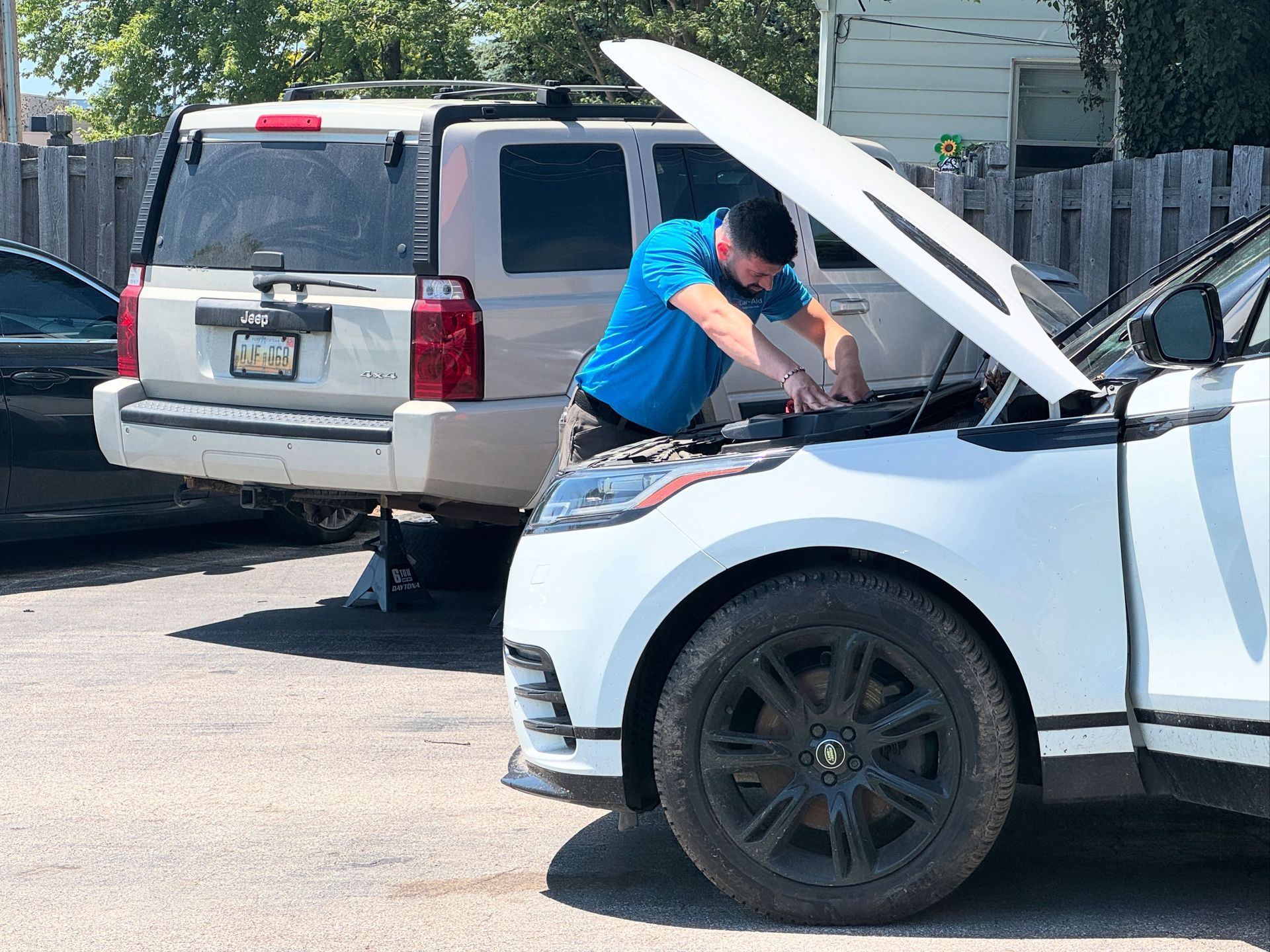 Mechanic works on a white car with the hood open, other vehicles parked behind him outdoors.