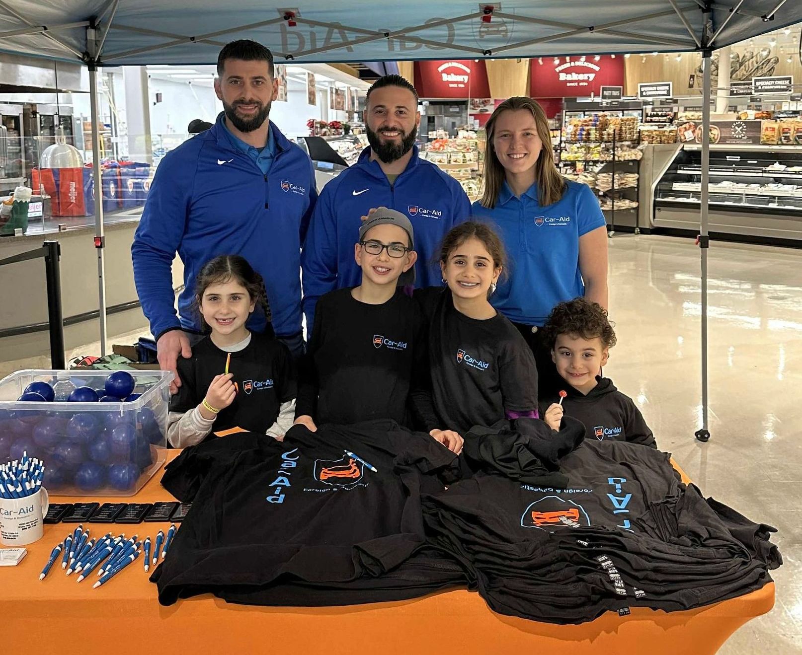 Group of adults and children at a promotional booth with blue and black shirts, pens, and lollipops inside a store.