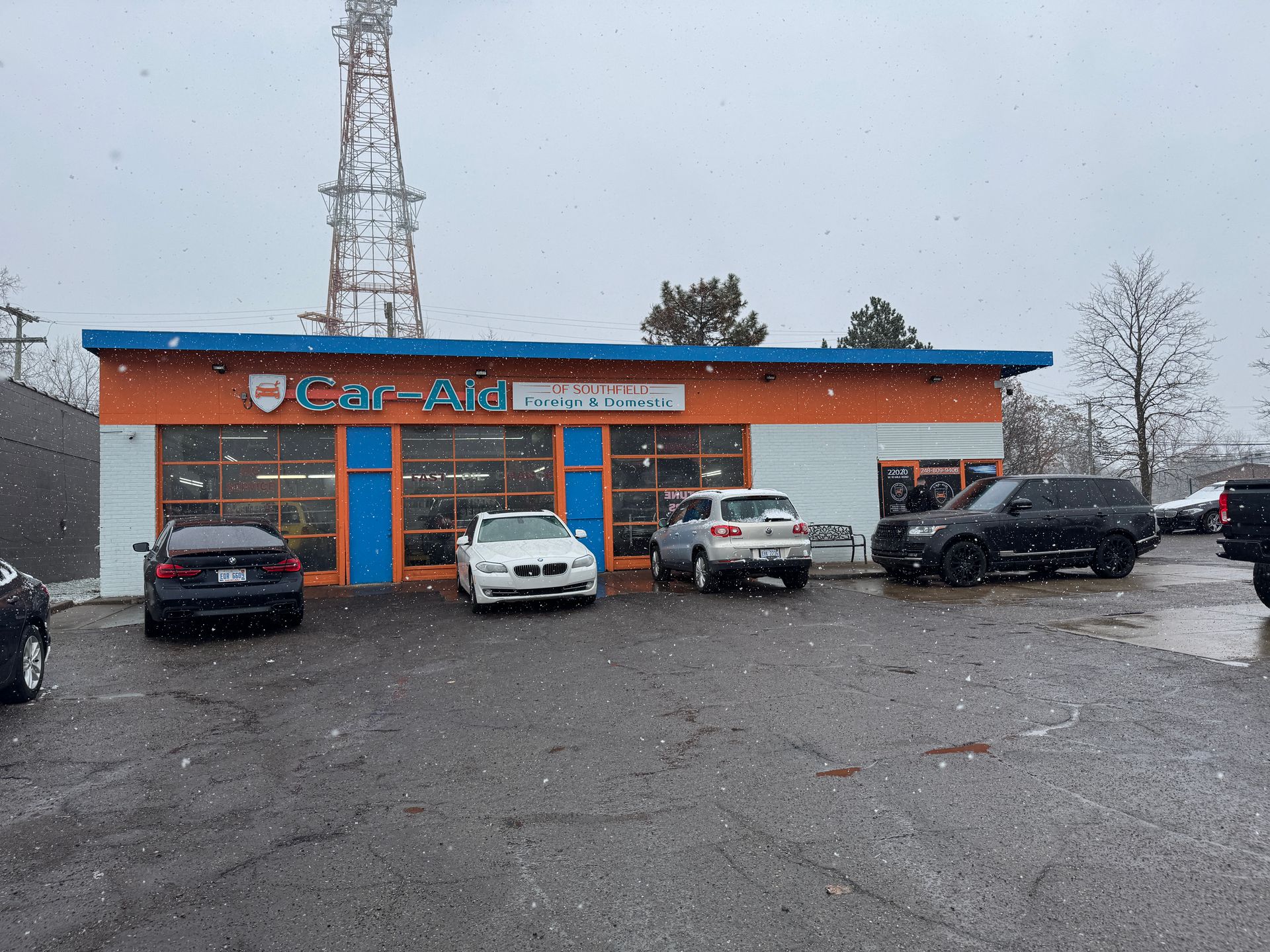 Cars parked outside a car repair shop with an orange facade, under snowy conditions. A telecommunications tower visible.