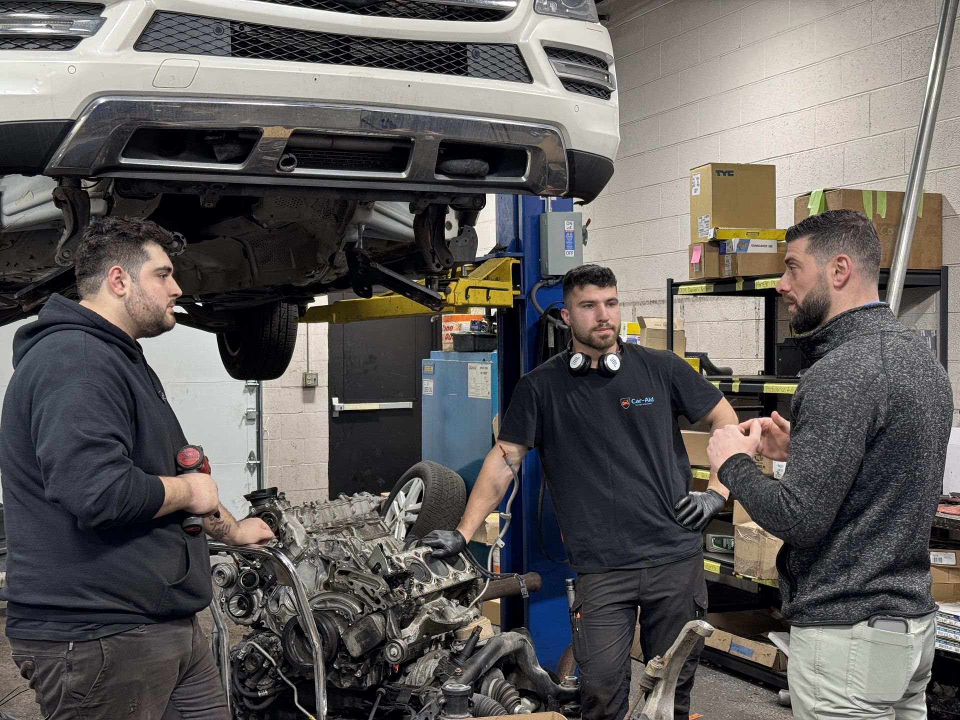 Three men are standing around a car engine in a garage.