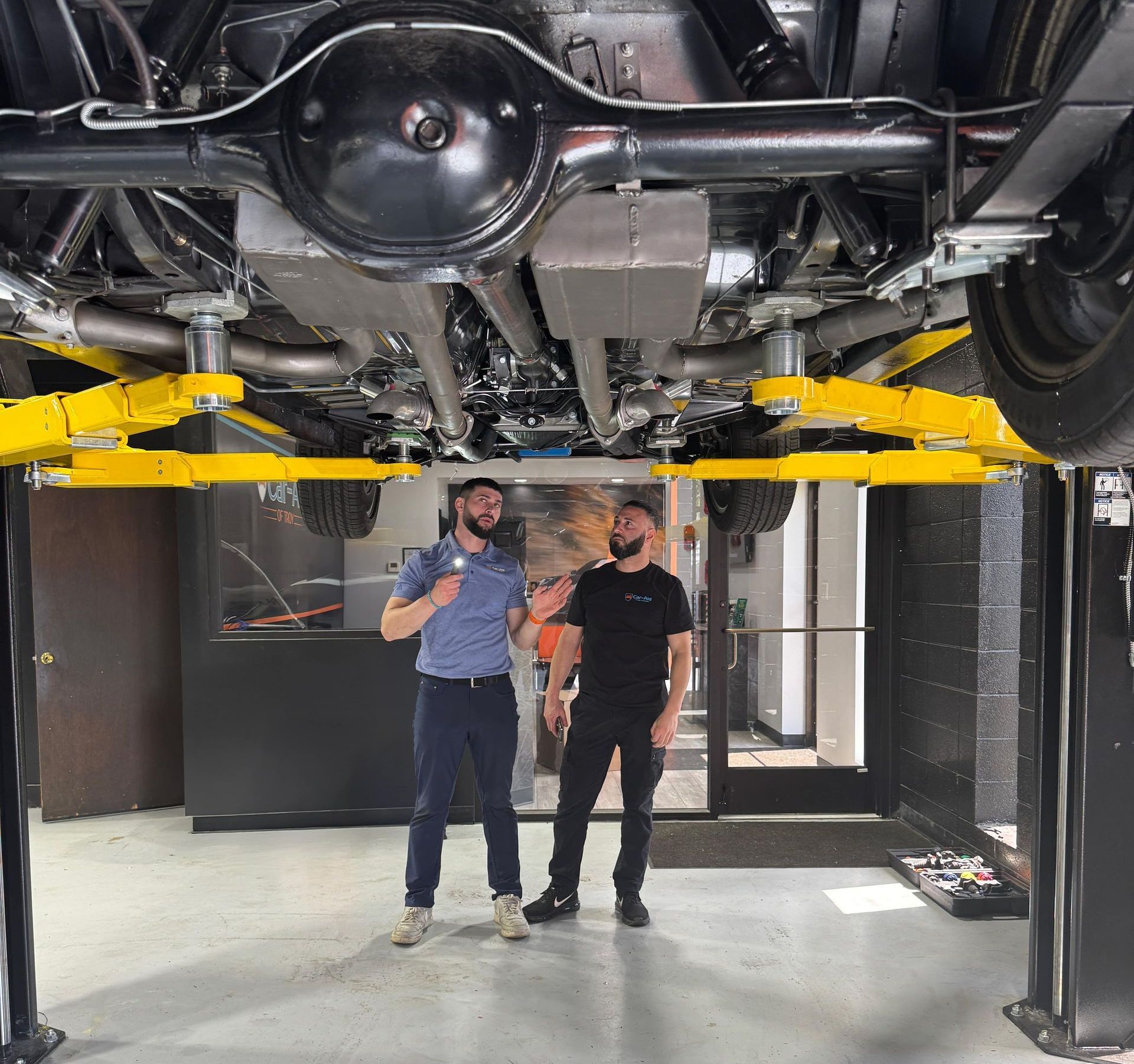Two men are standing under a car on a lift