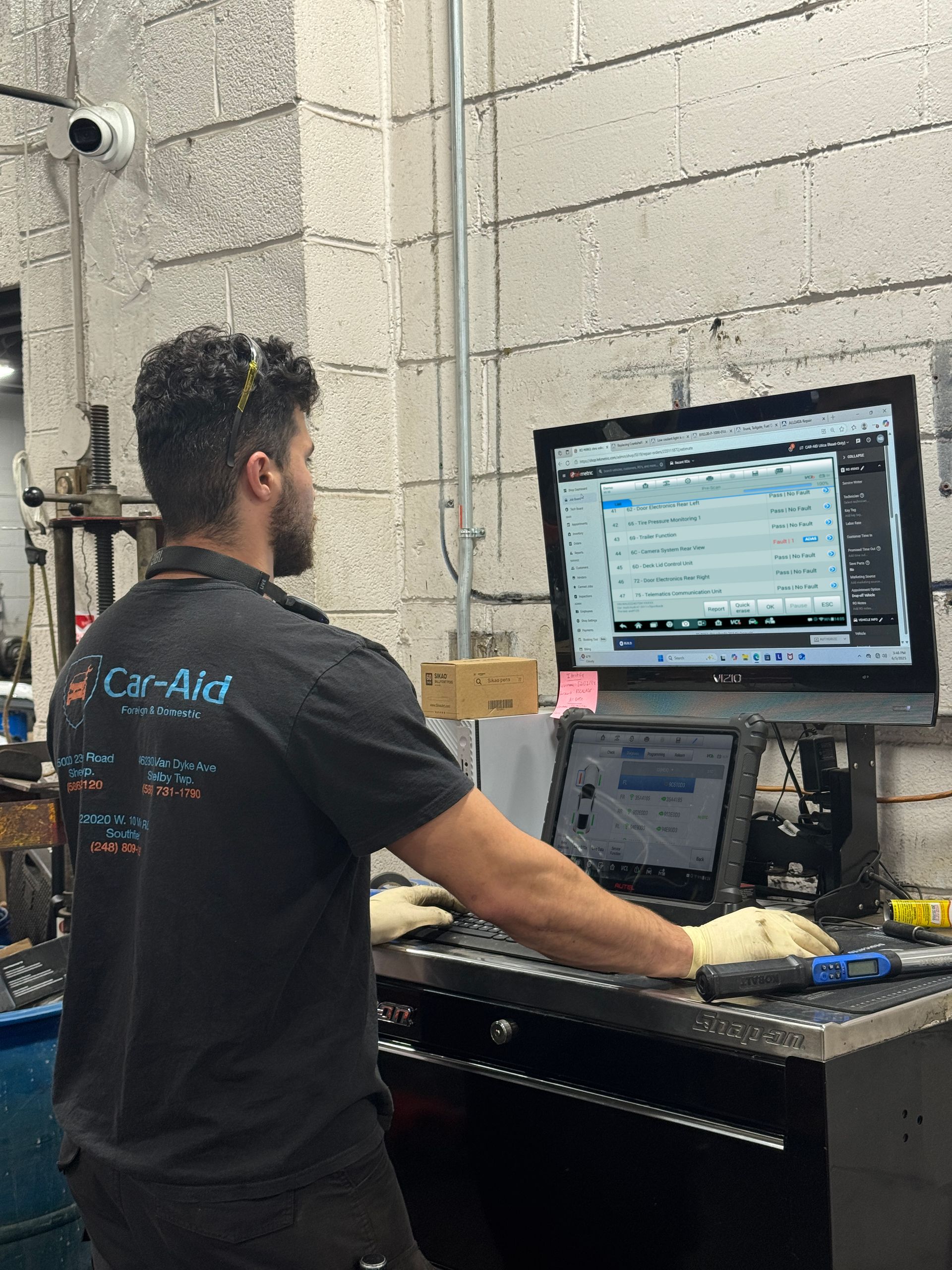 A mechanic in a shop uses a computer and tablet to diagnose a vehicle. He wears gloves and safety glasses, working near tools and equipment.