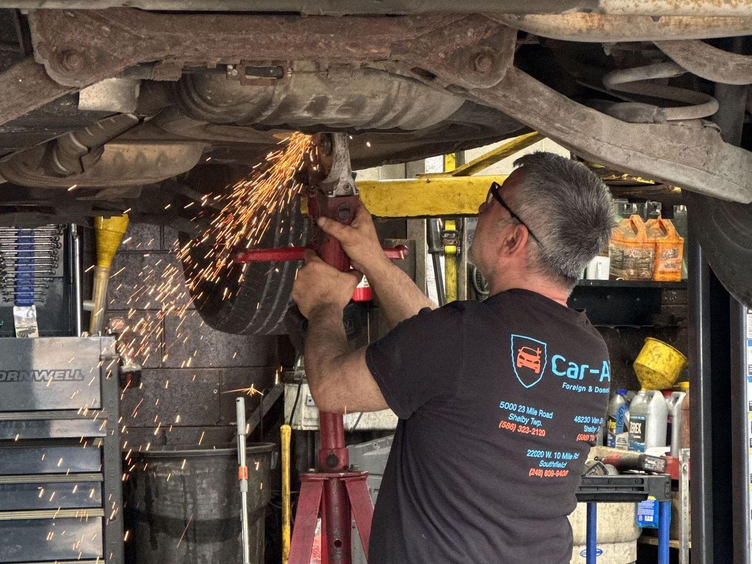 Mechanic using a grinder on the underside of a car, sparks flying. Shop interior with tools visible.