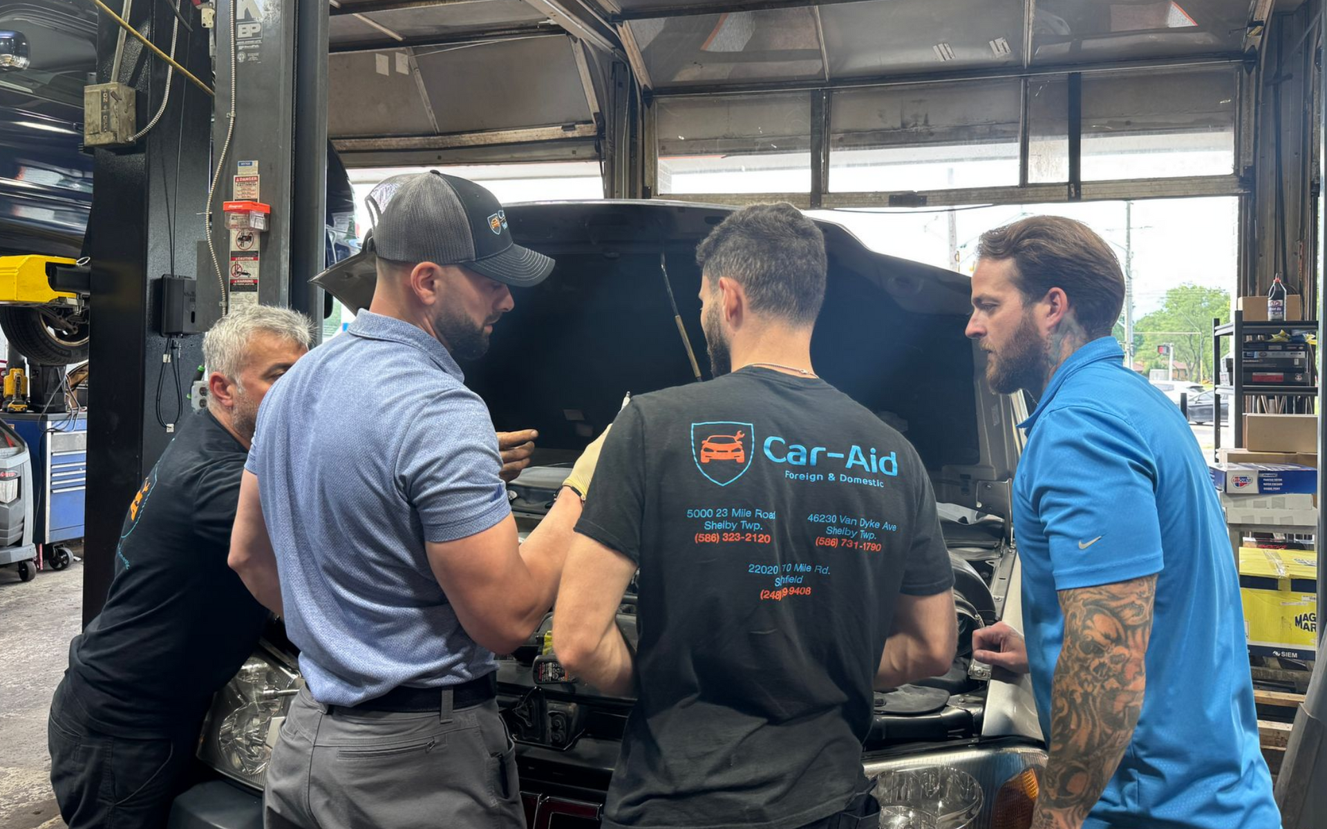 Four men examining a car engine in a garage. One wears a