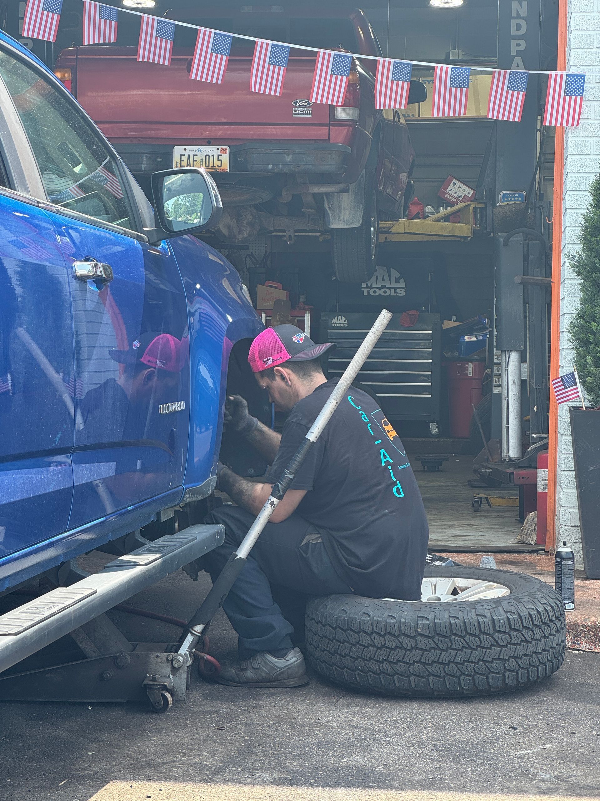 Mechanic changing tire on a blue truck in a garage, using a jack and sitting on a tire.